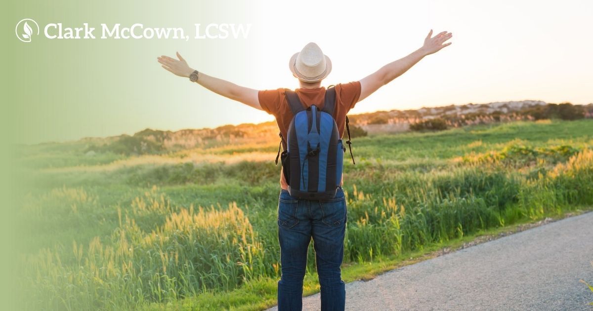 Person with backpack and hat stands on roadside with arms raised in a green field at sunset
