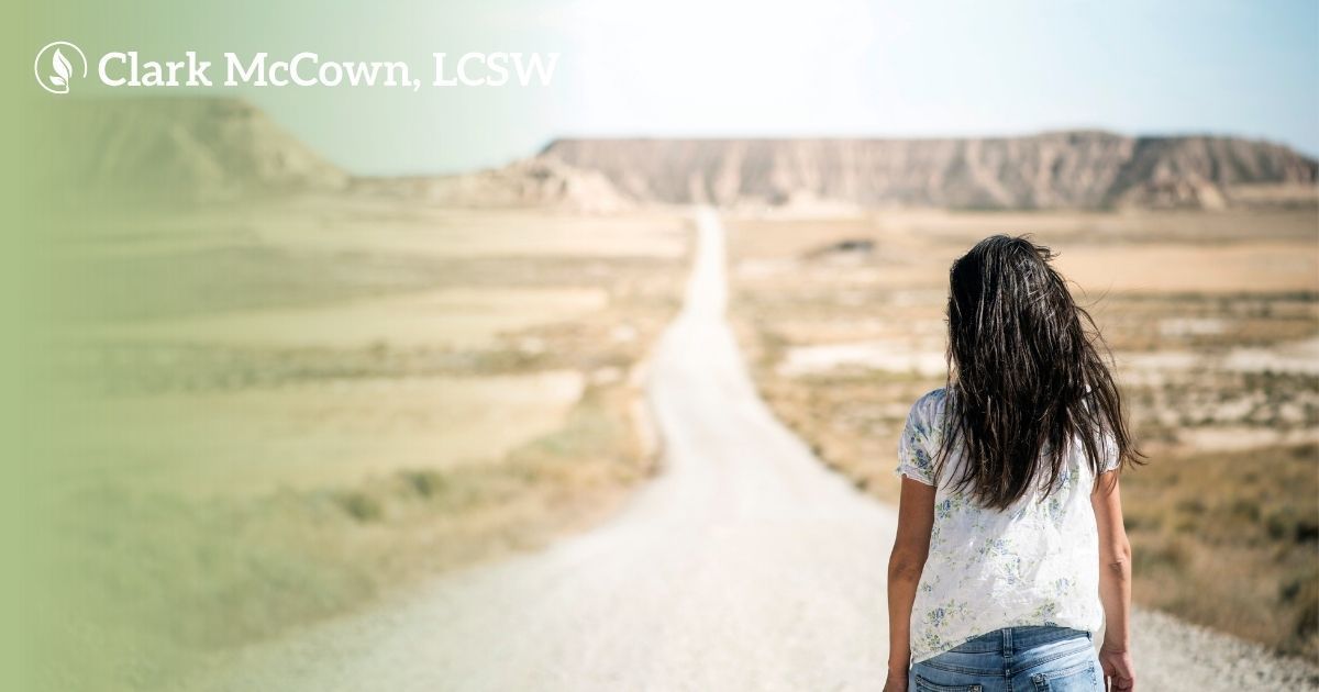 Woman with curly hair stands on a dirt road overlooking a wide, open landscape.