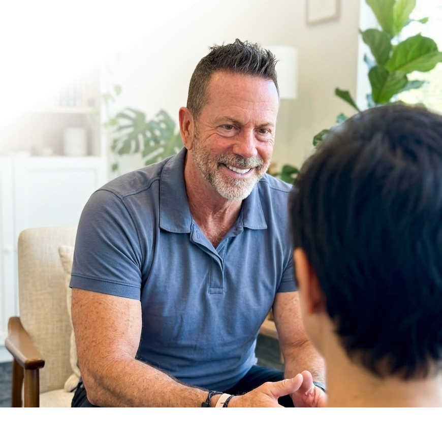 Two people talking in a bright living room, one smiling and leaning forward.