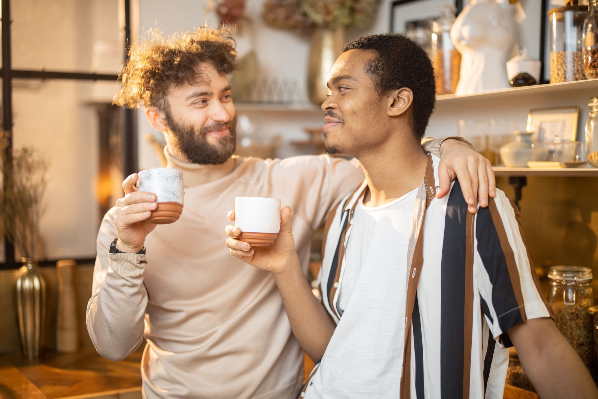 Two friends smiling and holding mugs in a cozy, sunlit kitchen