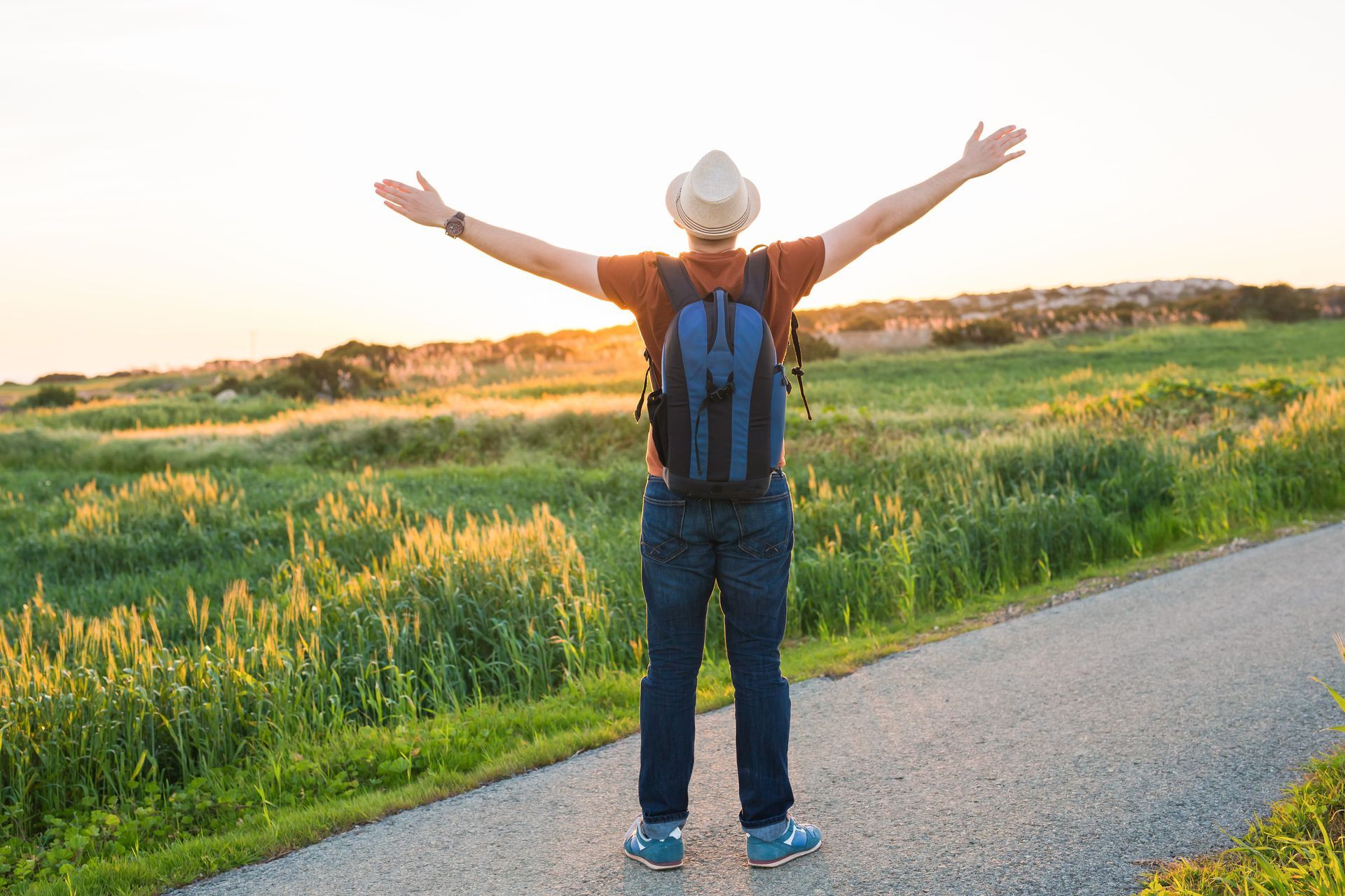 Person with backpack standing on a road with arms raised, overlooking a green field at sunset