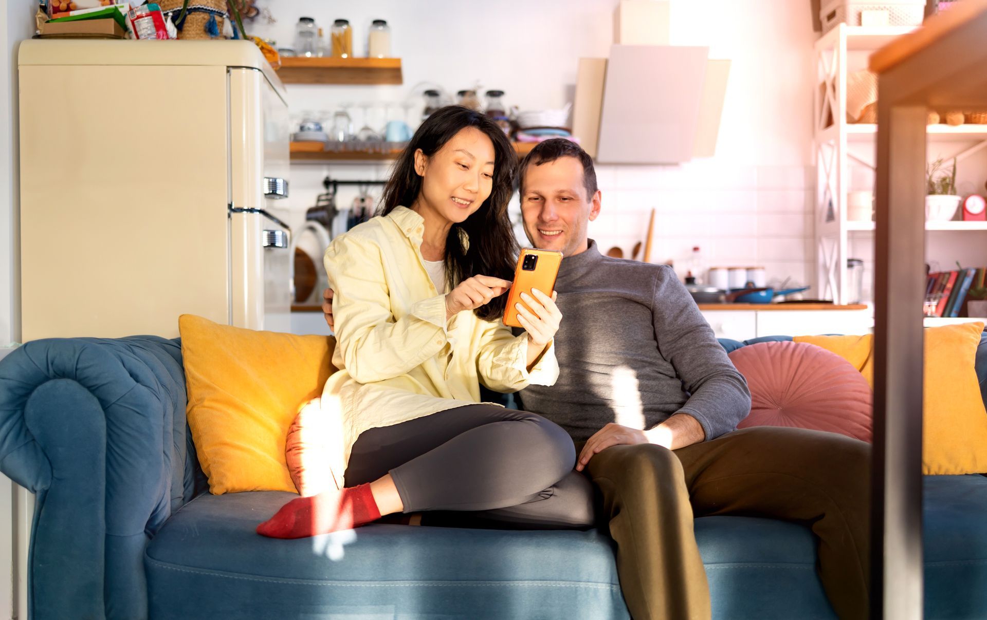 Two people sit on a couch, smiling at a smartphone in a cozy living room.