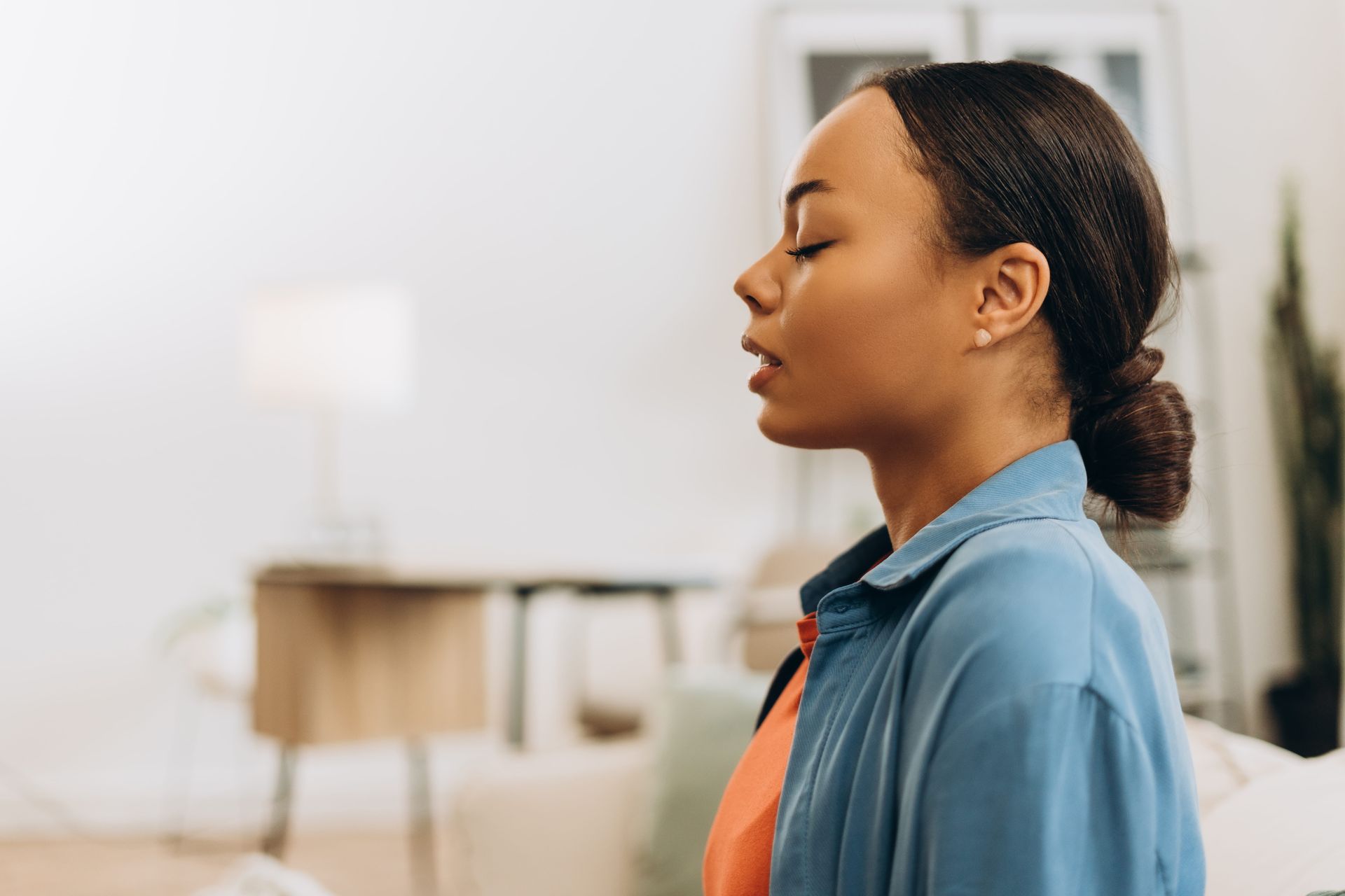Woman in a blue shirt standing in profile with eyes closed in a softly lit room