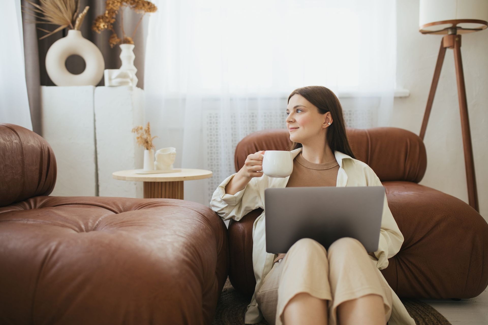 Woman on a sofa using a laptop and holding a mug in a bright living room