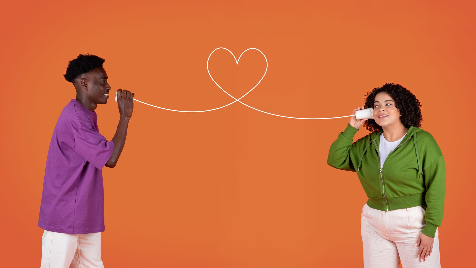 Two people in purple and green shirts blowing a dashed line that forms a heart on an orange background.
