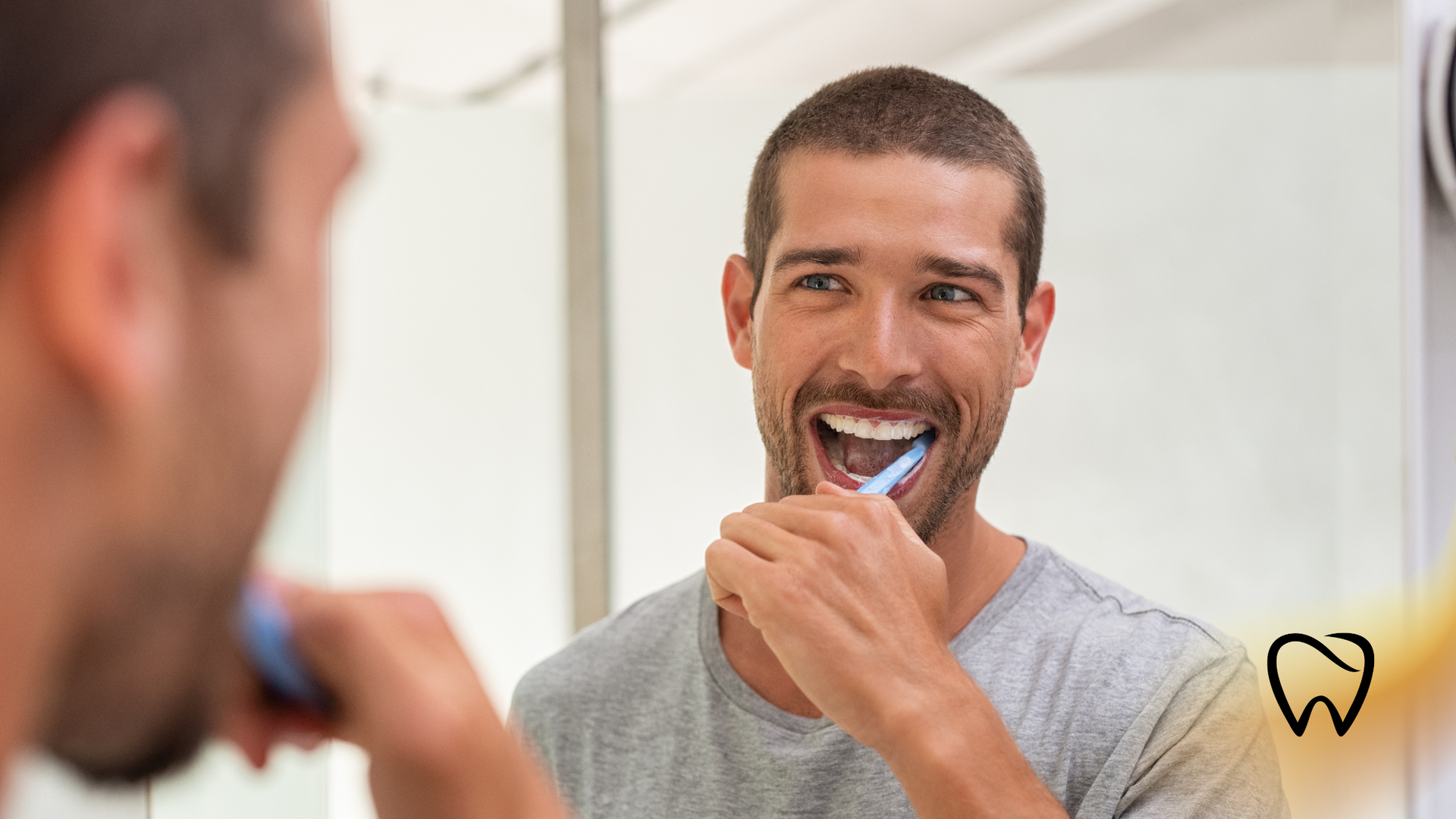 Man brushing teeth in front of a mirror, smiling, gray shirt, bathroom.