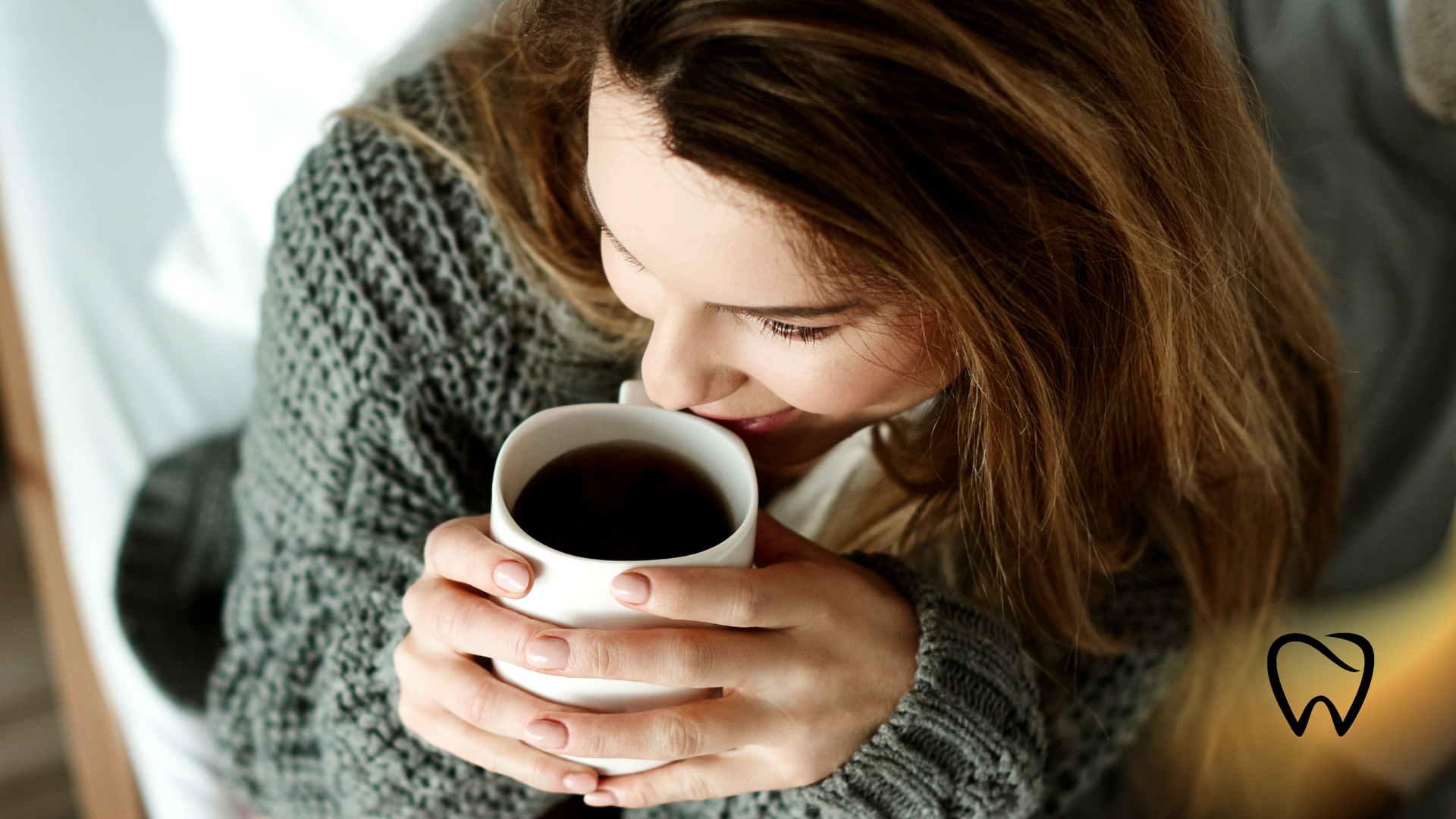 Woman in gray sweater, sipping from a white mug, eyes closed, indoors.