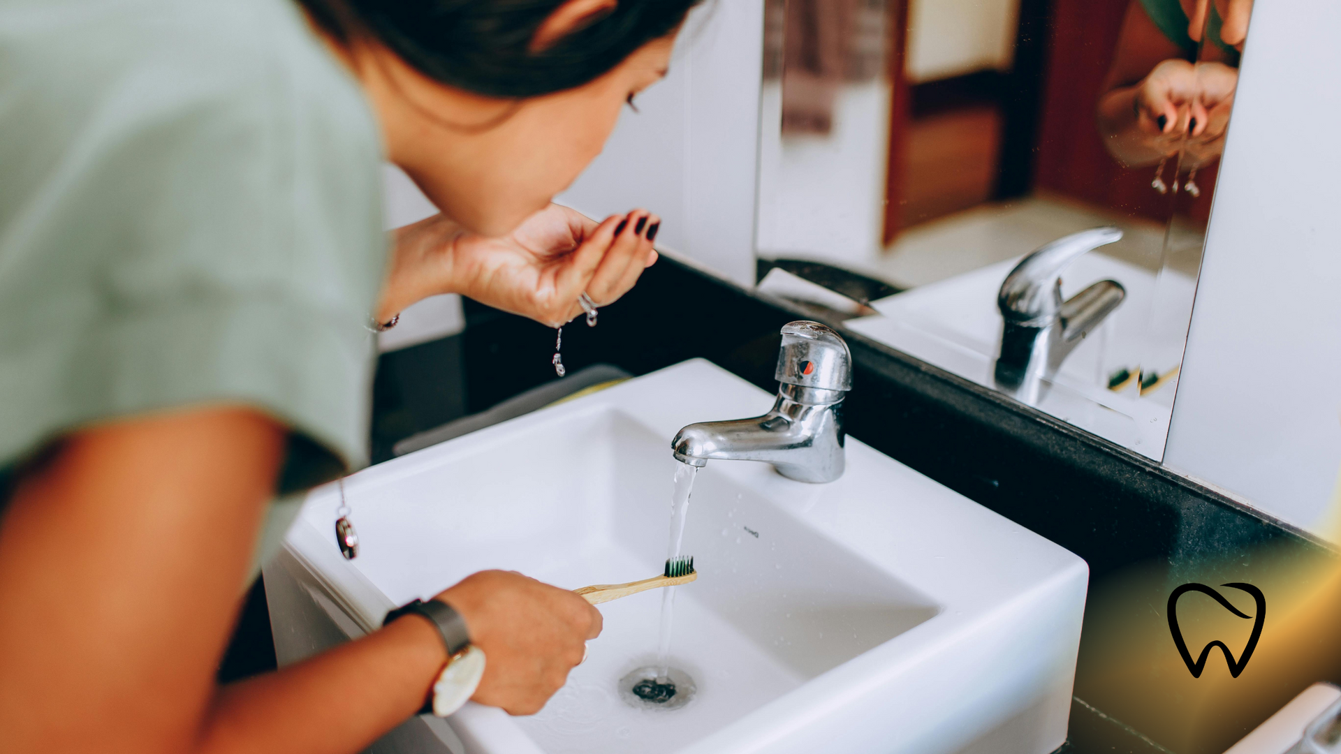 Woman rinsing face in a bathroom sink, holding a toothbrush. Reflection visible in mirror.