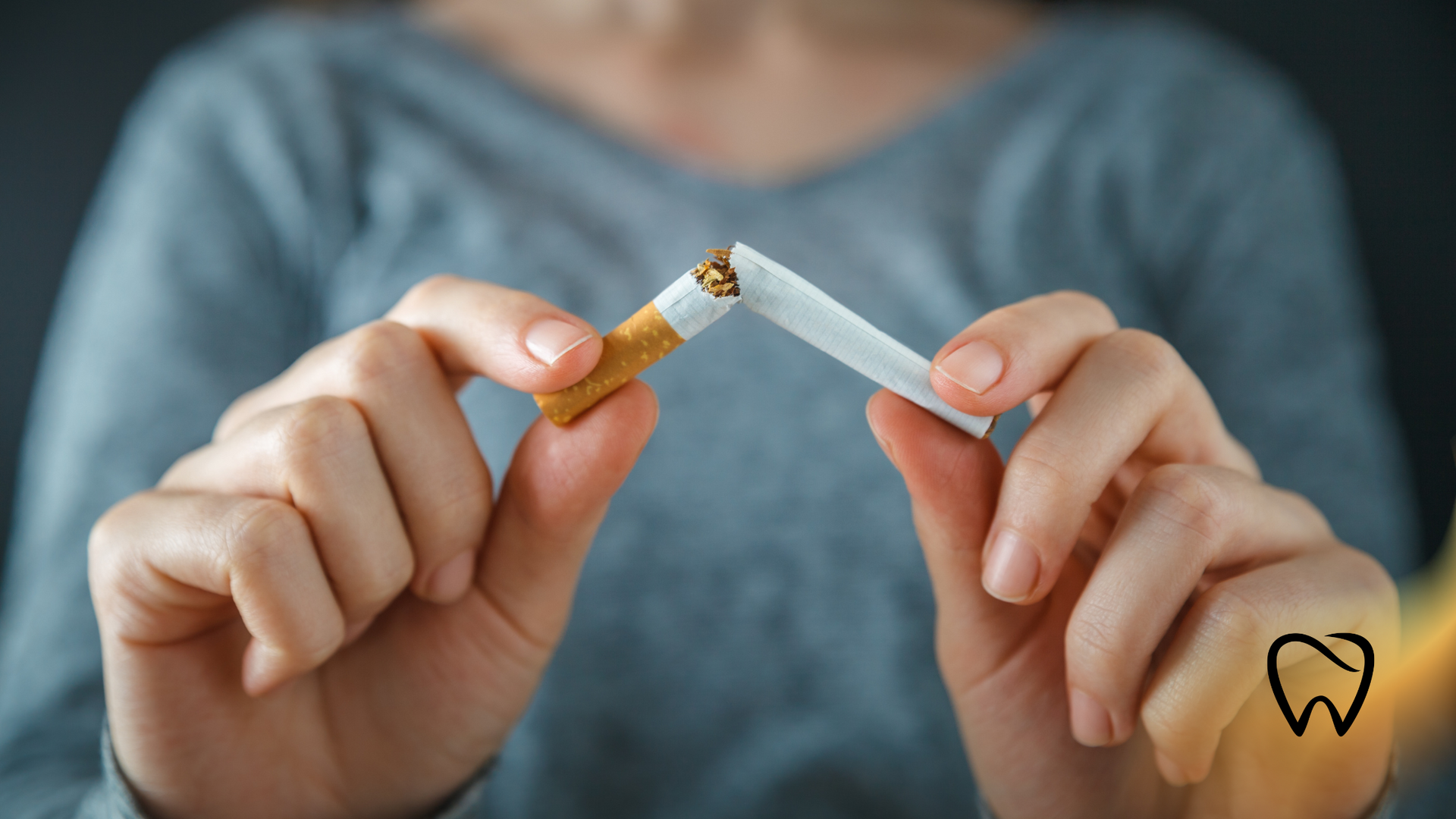 Person breaking a cigarette in half; gray shirt and dark background.