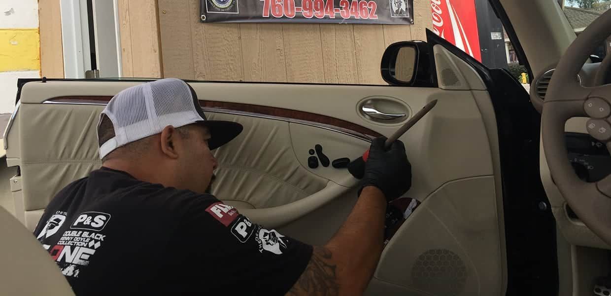 A man in a baseball cap works on a car door, likely repairing the upholstery, in a shop setting.