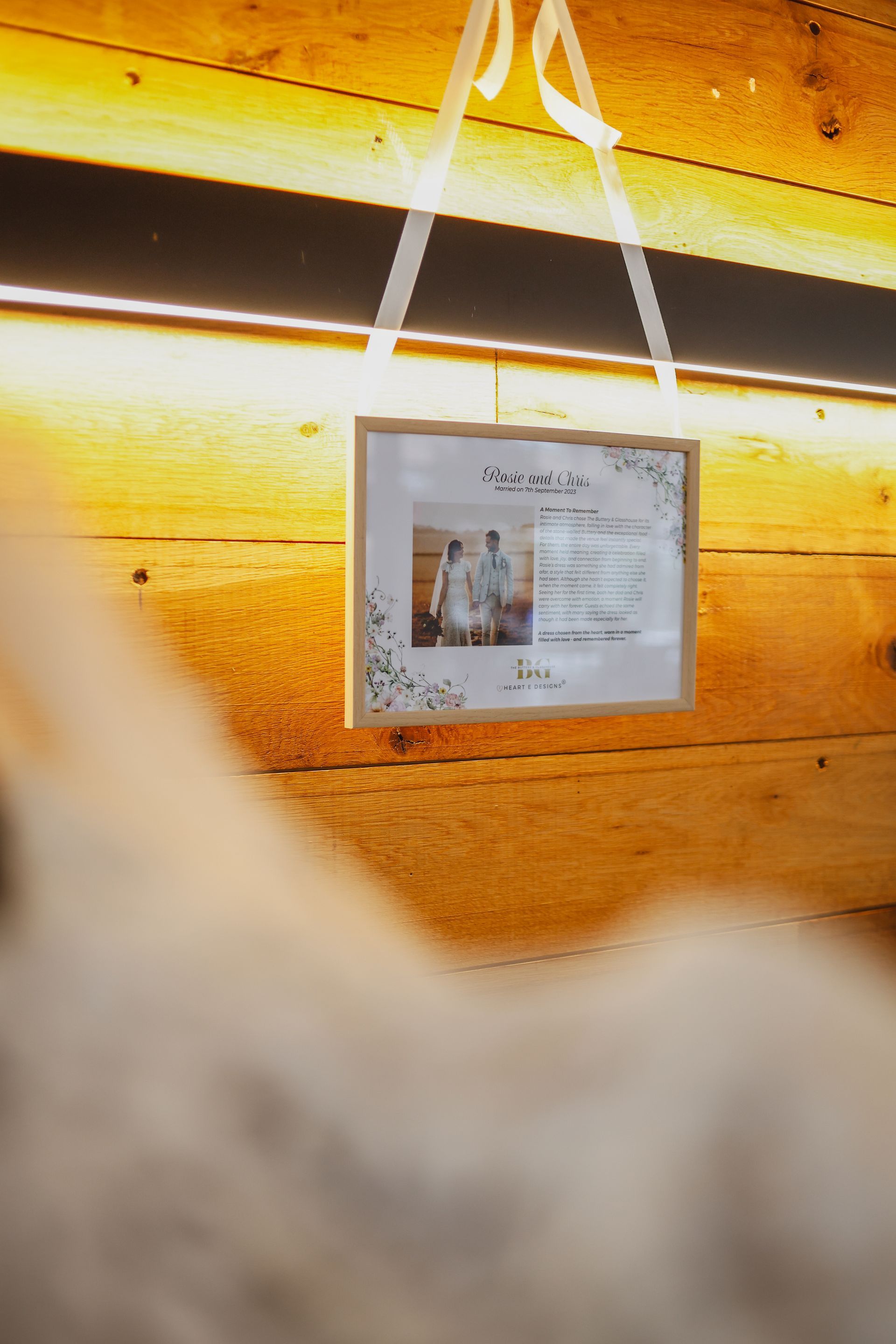 Framed photo hanging by white ribbon on a warm wooden wall, softly lit from above
