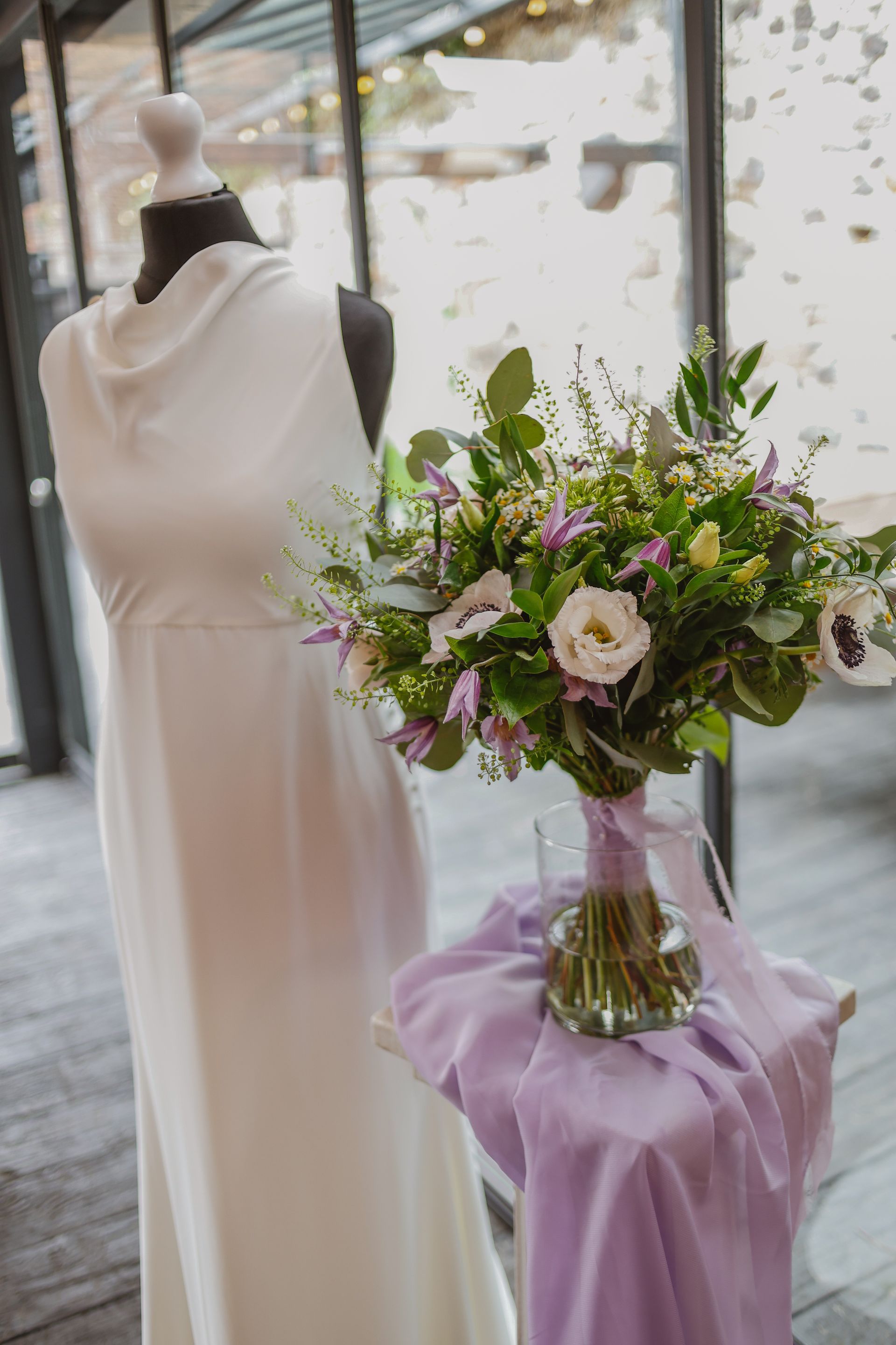 White dress on mannequin beside a lavender-draped table with a large pink-and-green floral arrangement