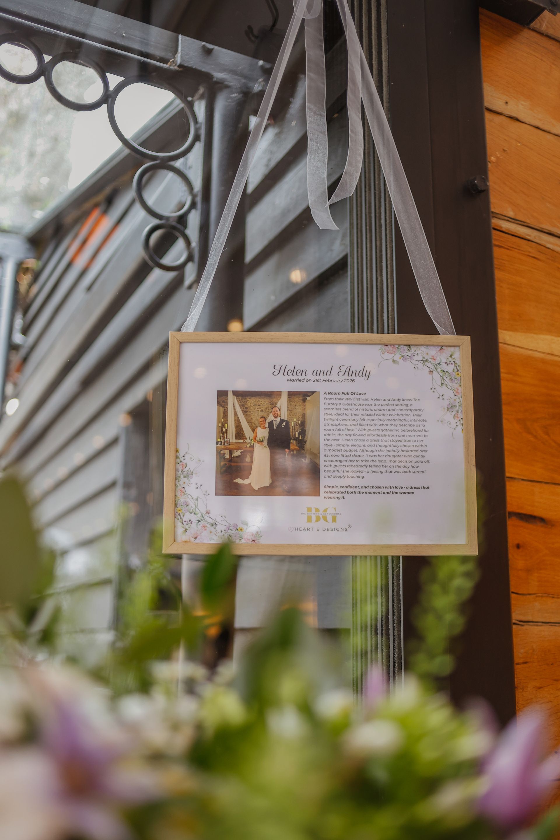 Framed sign hanging outside a wooden building, with greenery blurred in the foreground.
