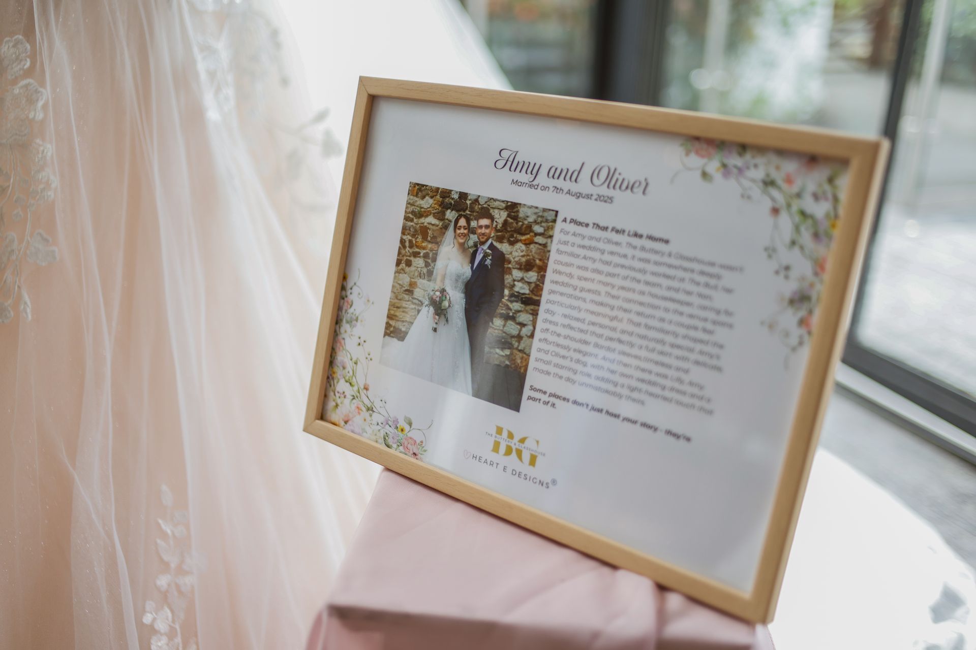 Framed wedding sign with photo and text on a soft pink table by a dress and window