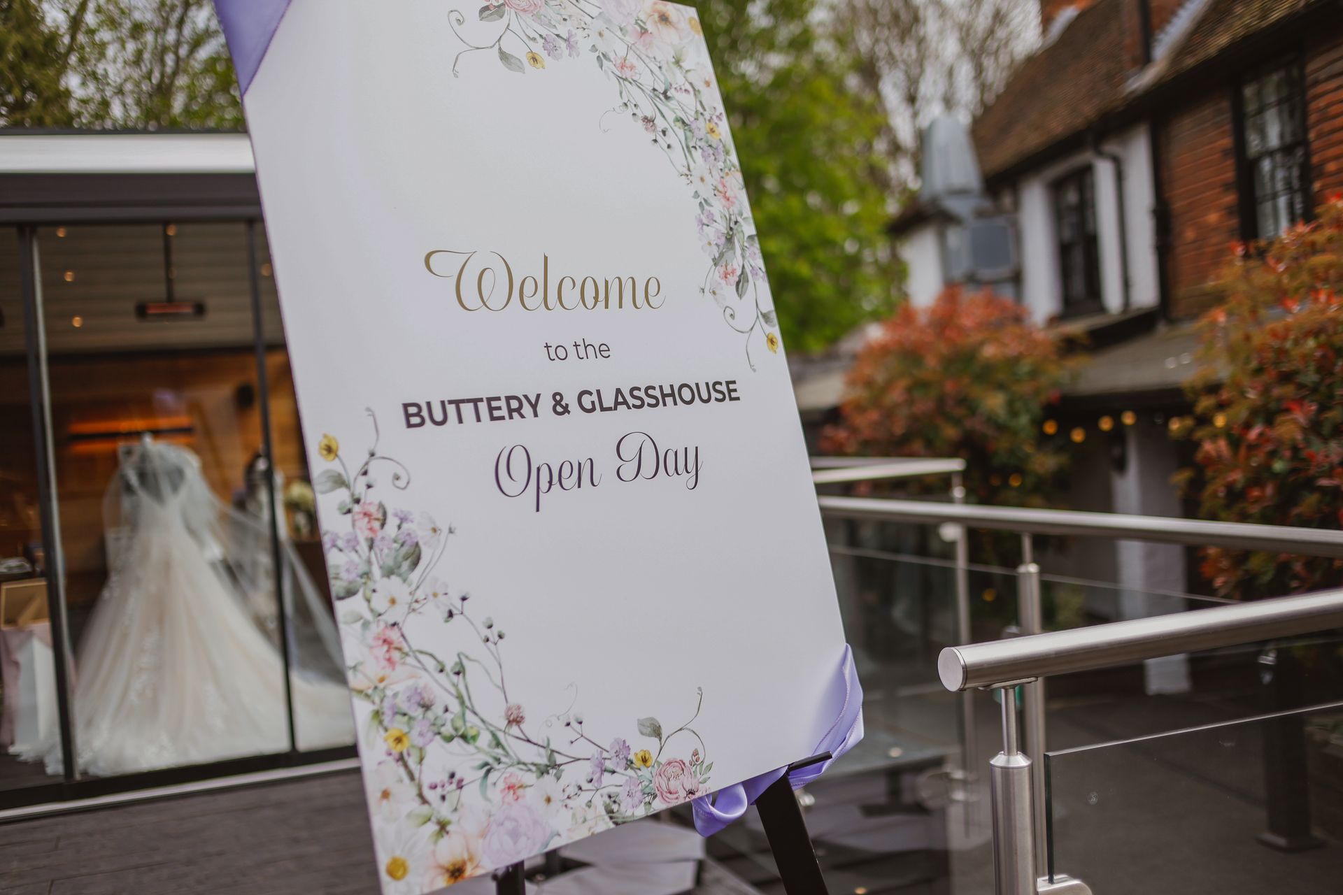 Wedding welcome sign on an outdoor terrace, with floral border and a dress in the background