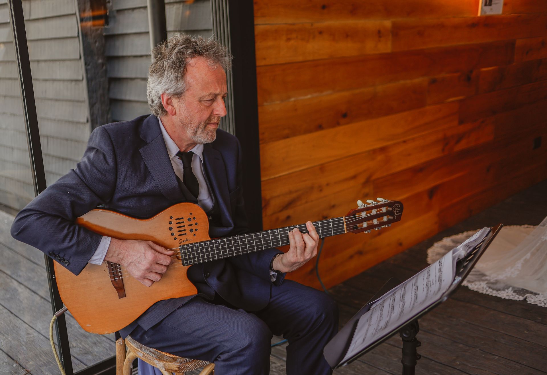 Man seated on a porch playing an acoustic guitar beside a music stand