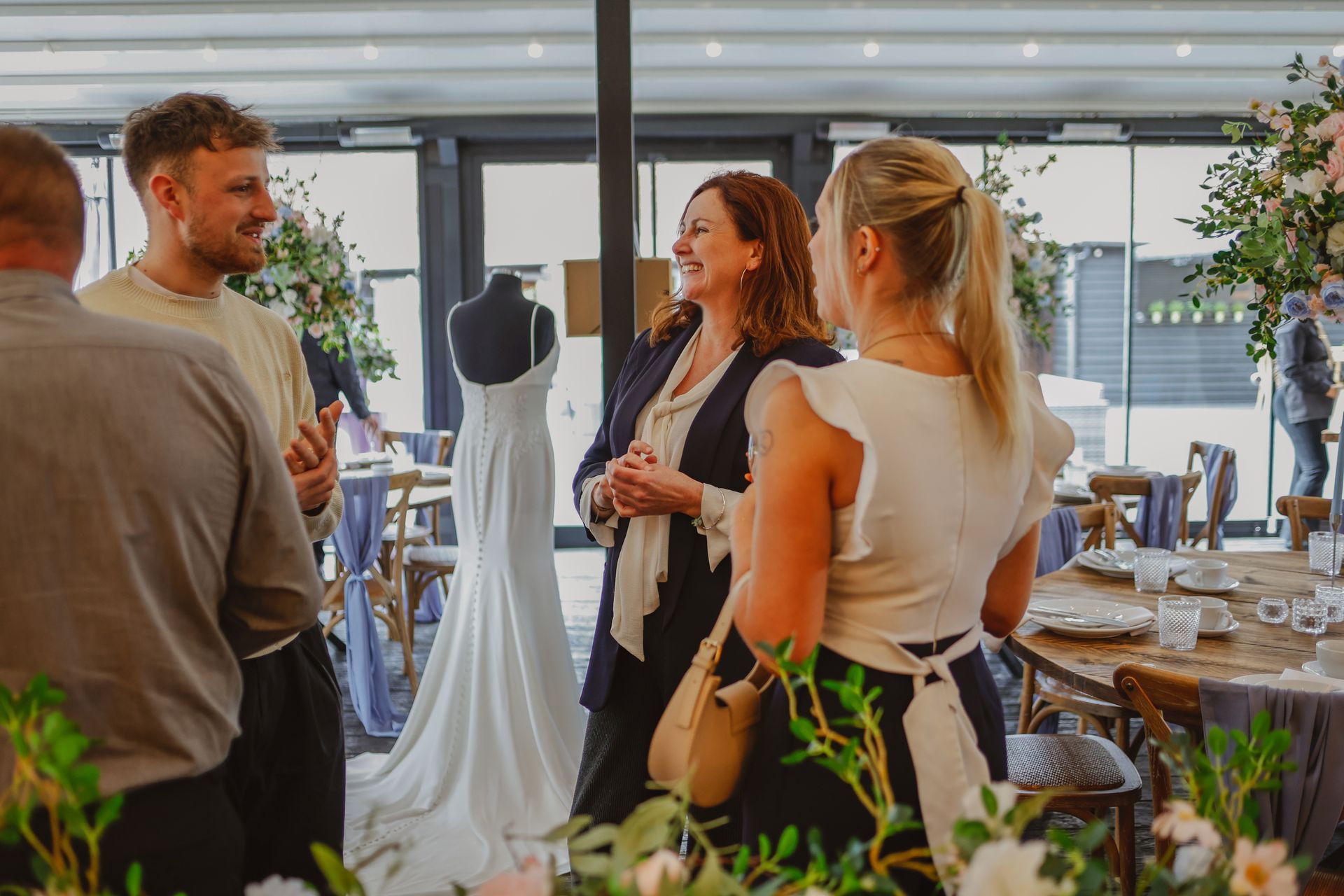 Guests chatting at a wedding reception with tables, flowers, and a white dress on display