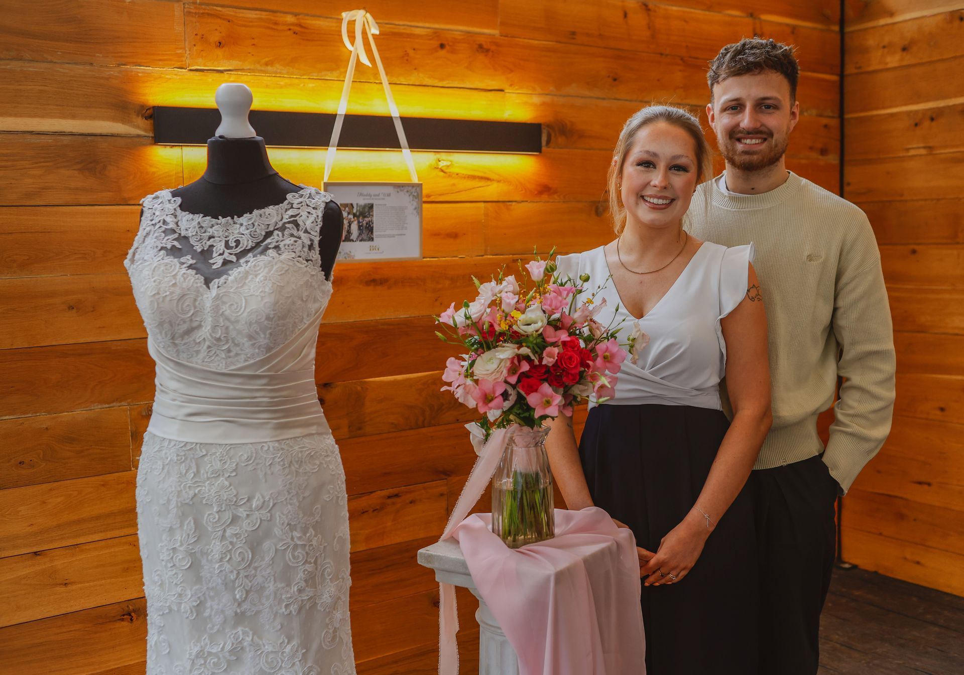Bride and groom standing beside a wedding dress and bouquet in a wooden room