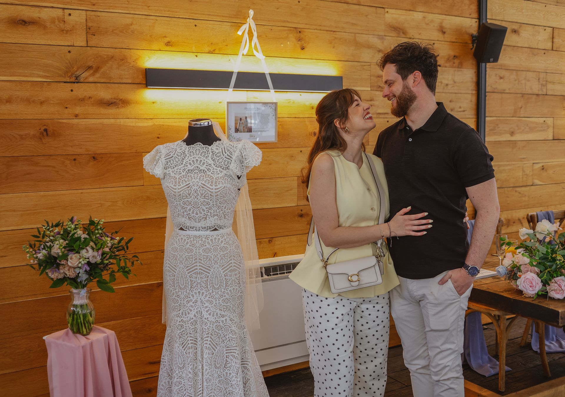 Woman and man standing close in a wood-paneled boutique beside a display dress and flowers