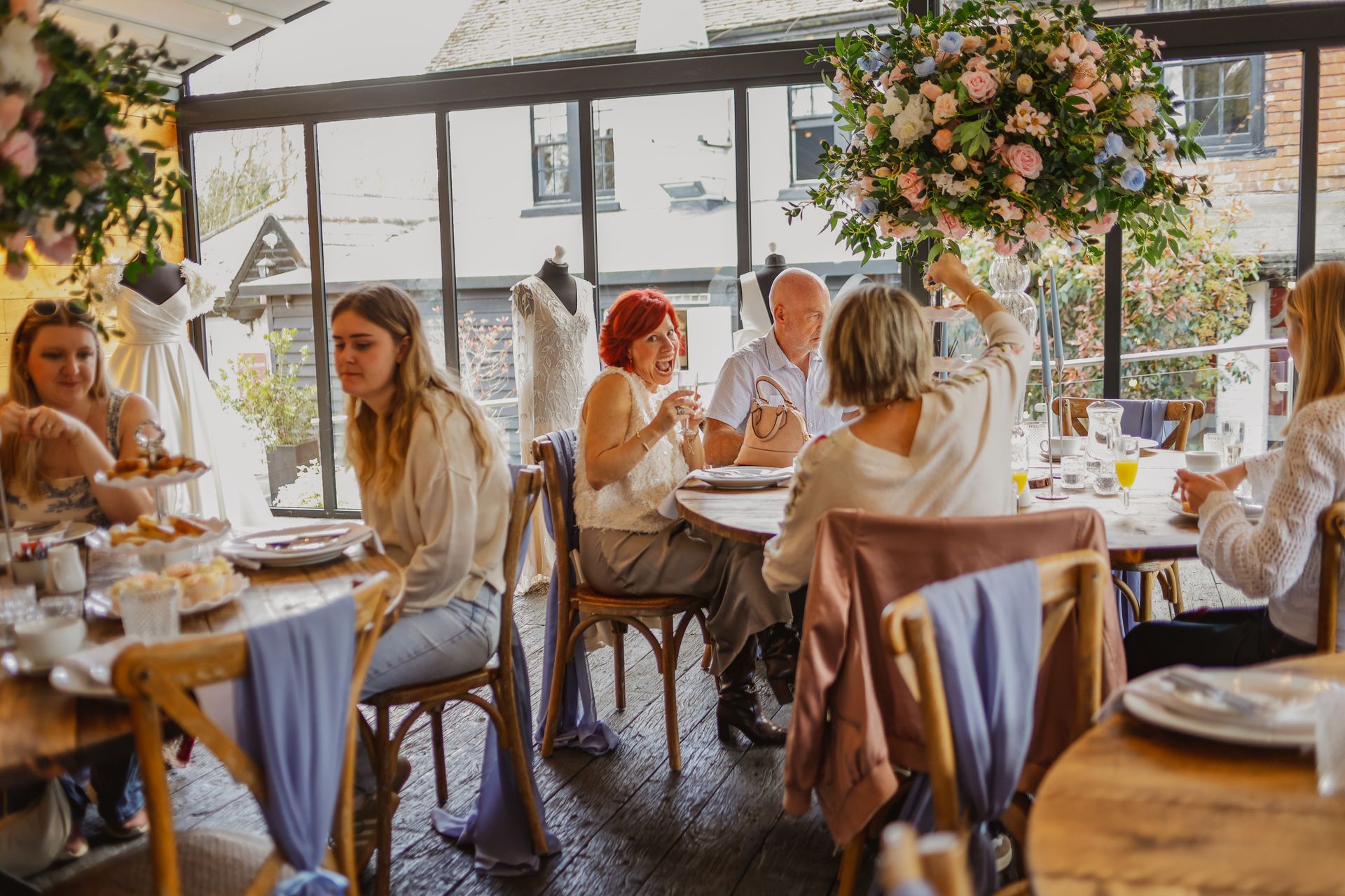 Guests dining at a warmly lit, flower-decorated café with large windows and white table settings