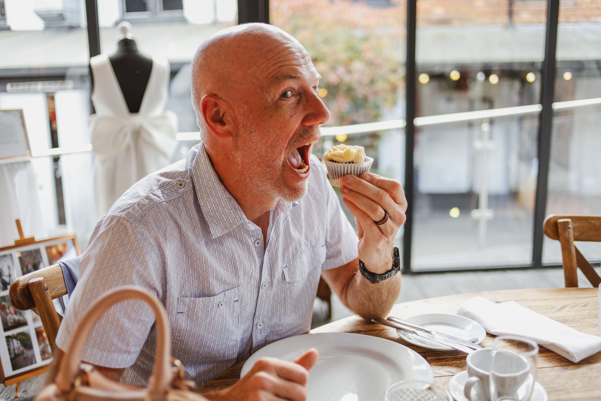Man seated at a table, eating a small pastry in a bright café by a window.