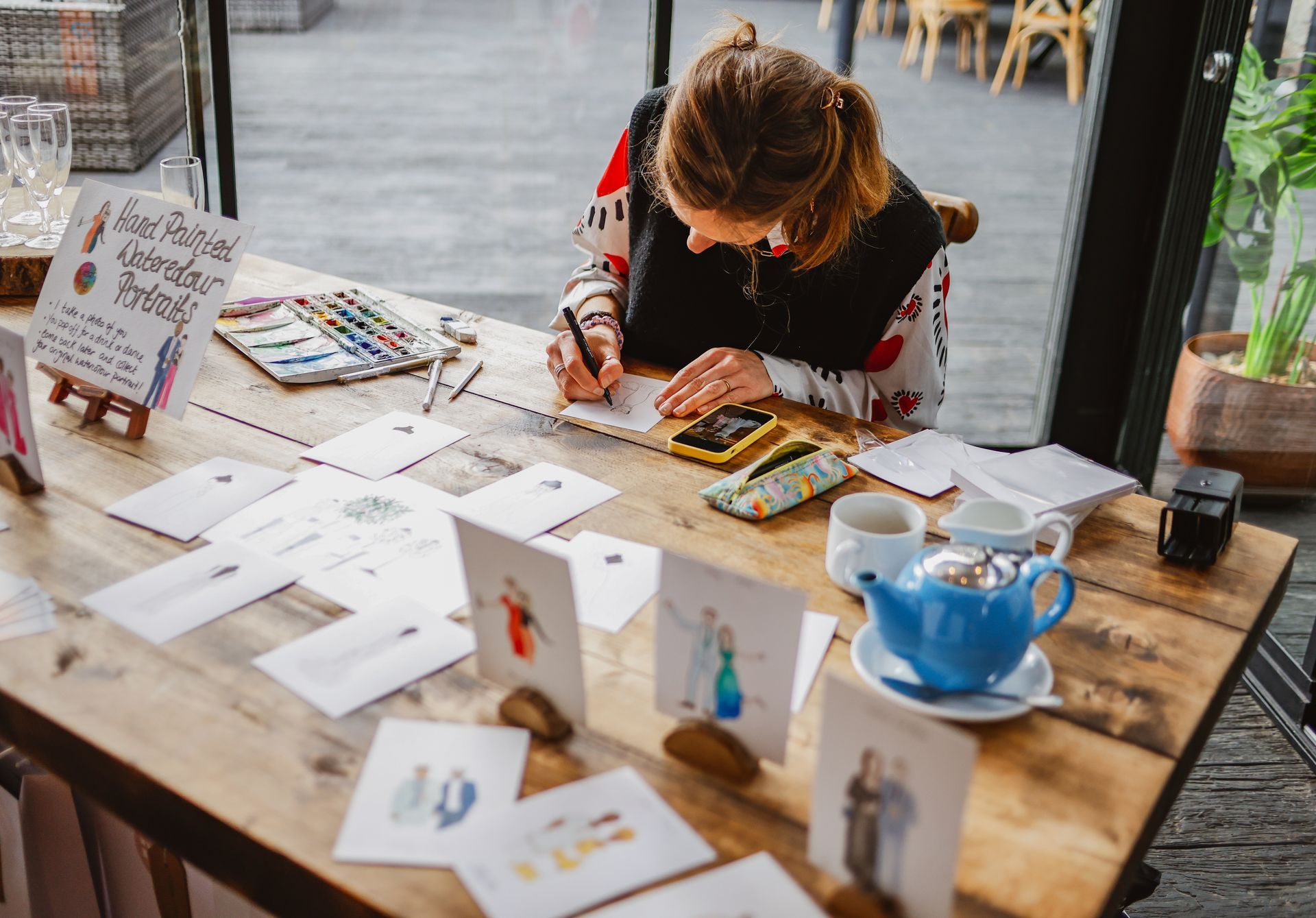Person painting at a café table with papers, postcards, and a blue teacup spread around.