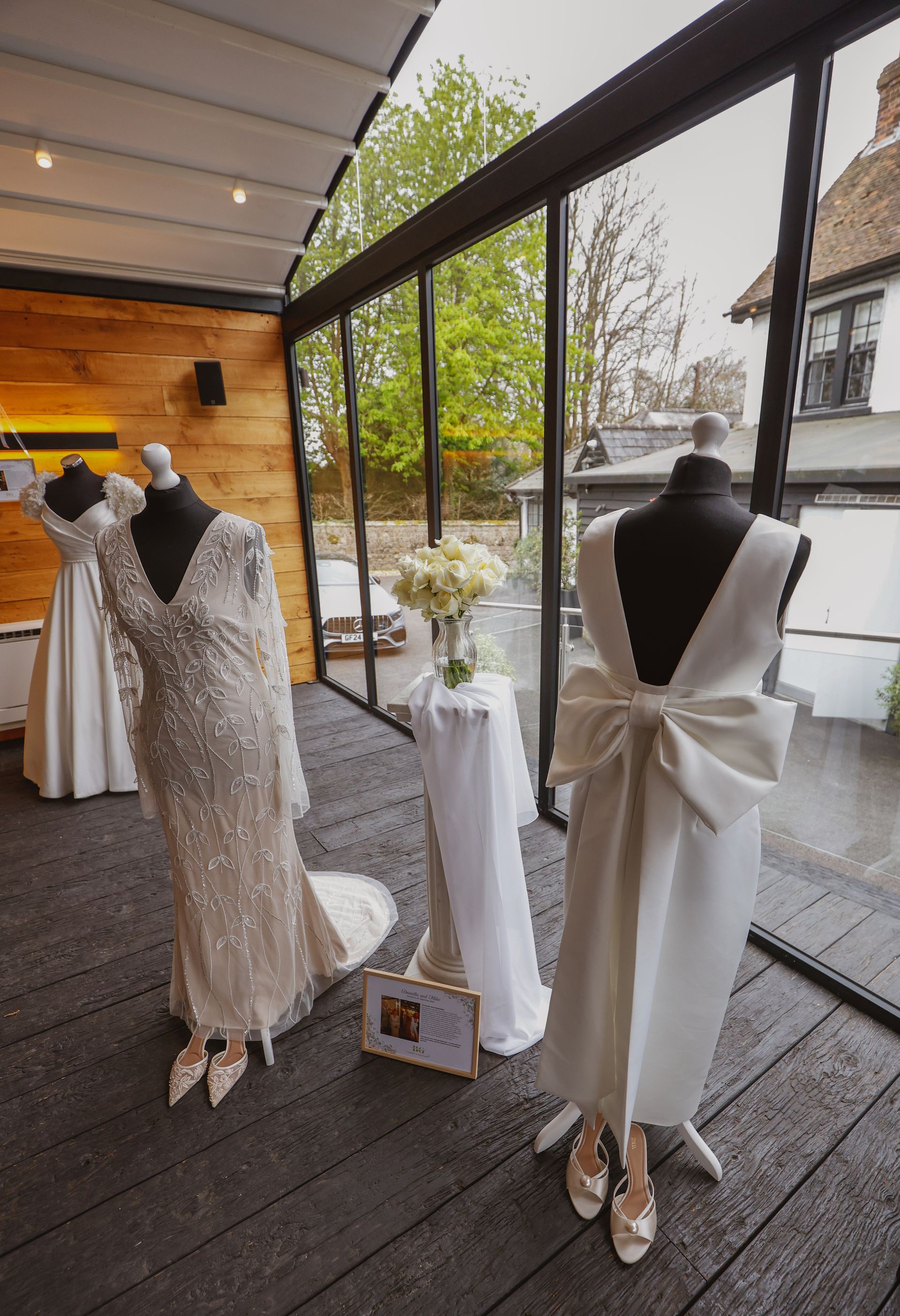 Wedding dresses on mannequins in a boutique window display, including a lace gown and a white dress with a bow.