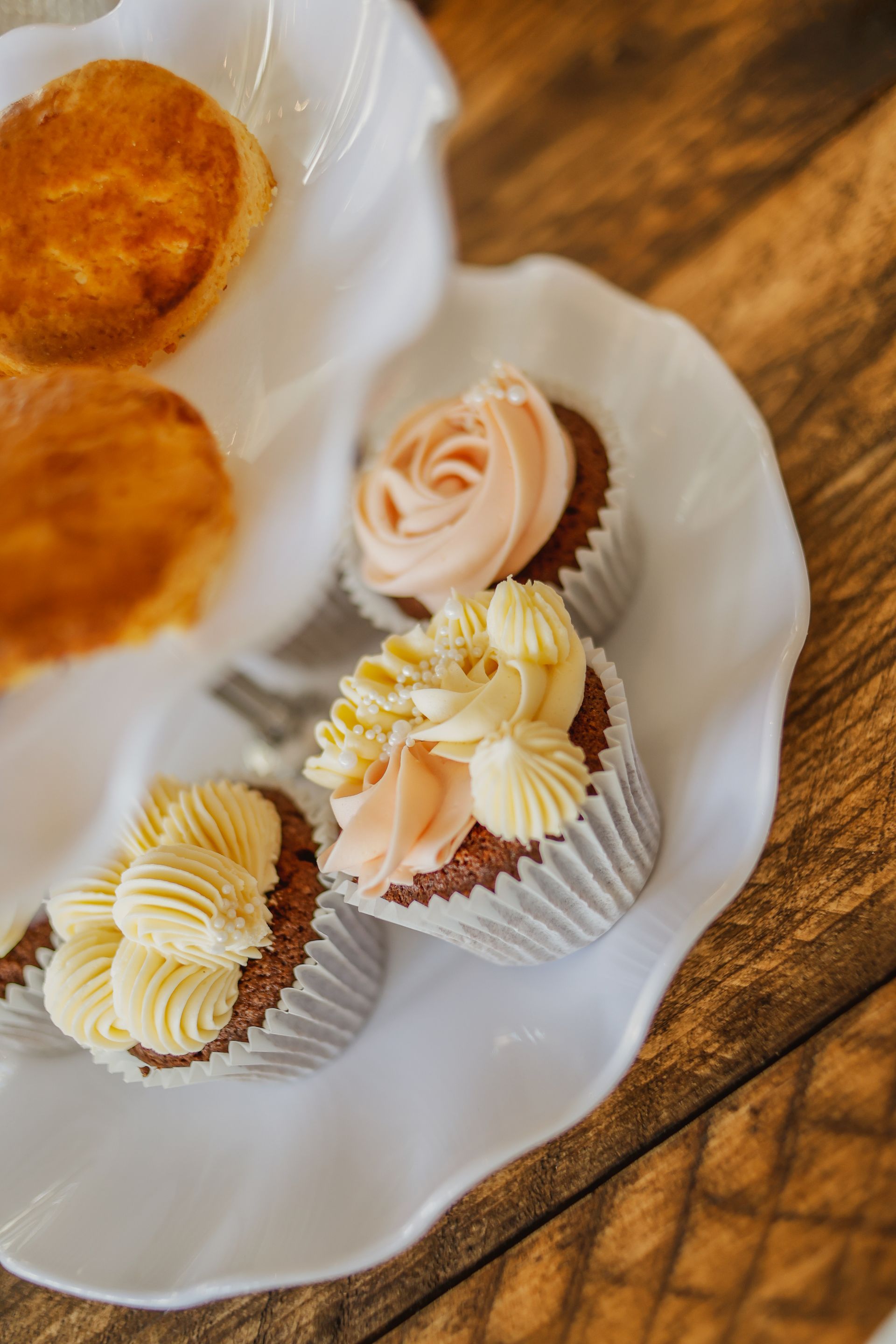 Three frosted cupcakes on a white plate beside two orange cookies on a wooden table