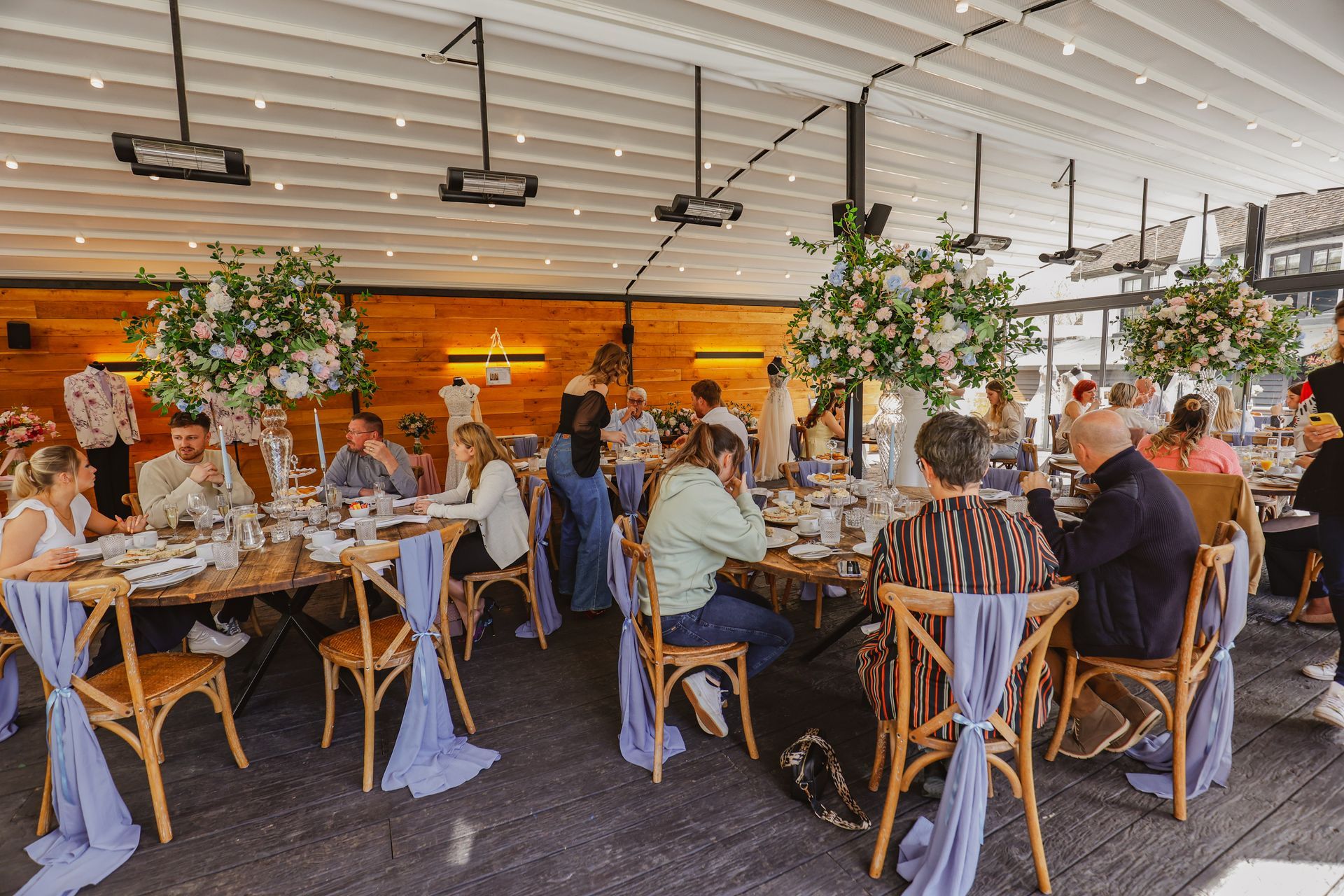 Guests dining at a bright outdoor restaurant with wooden tables and greenery under a white canopy