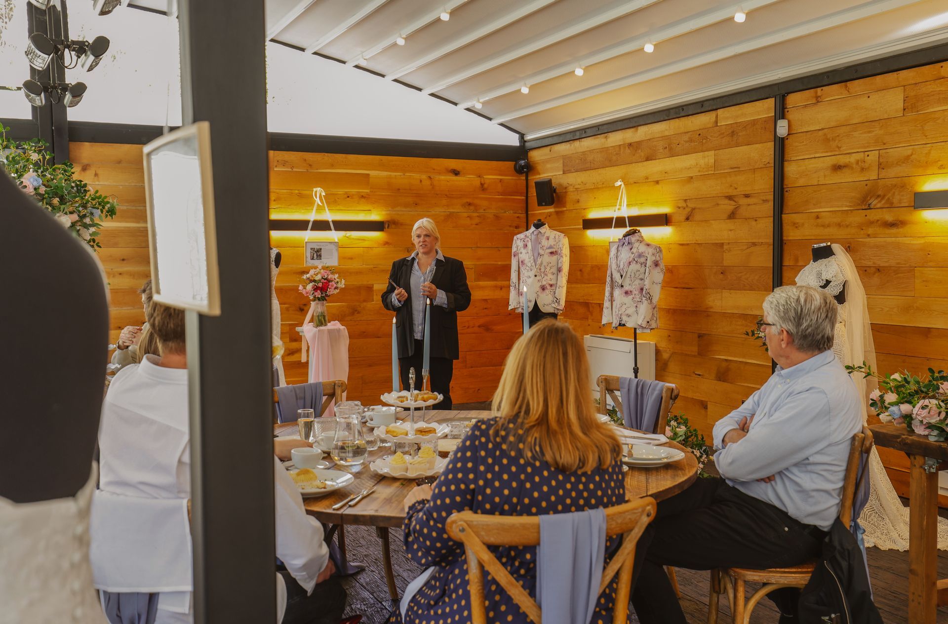 People seated at a table listen to a speaker presenting in a warmly lit indoor venue.