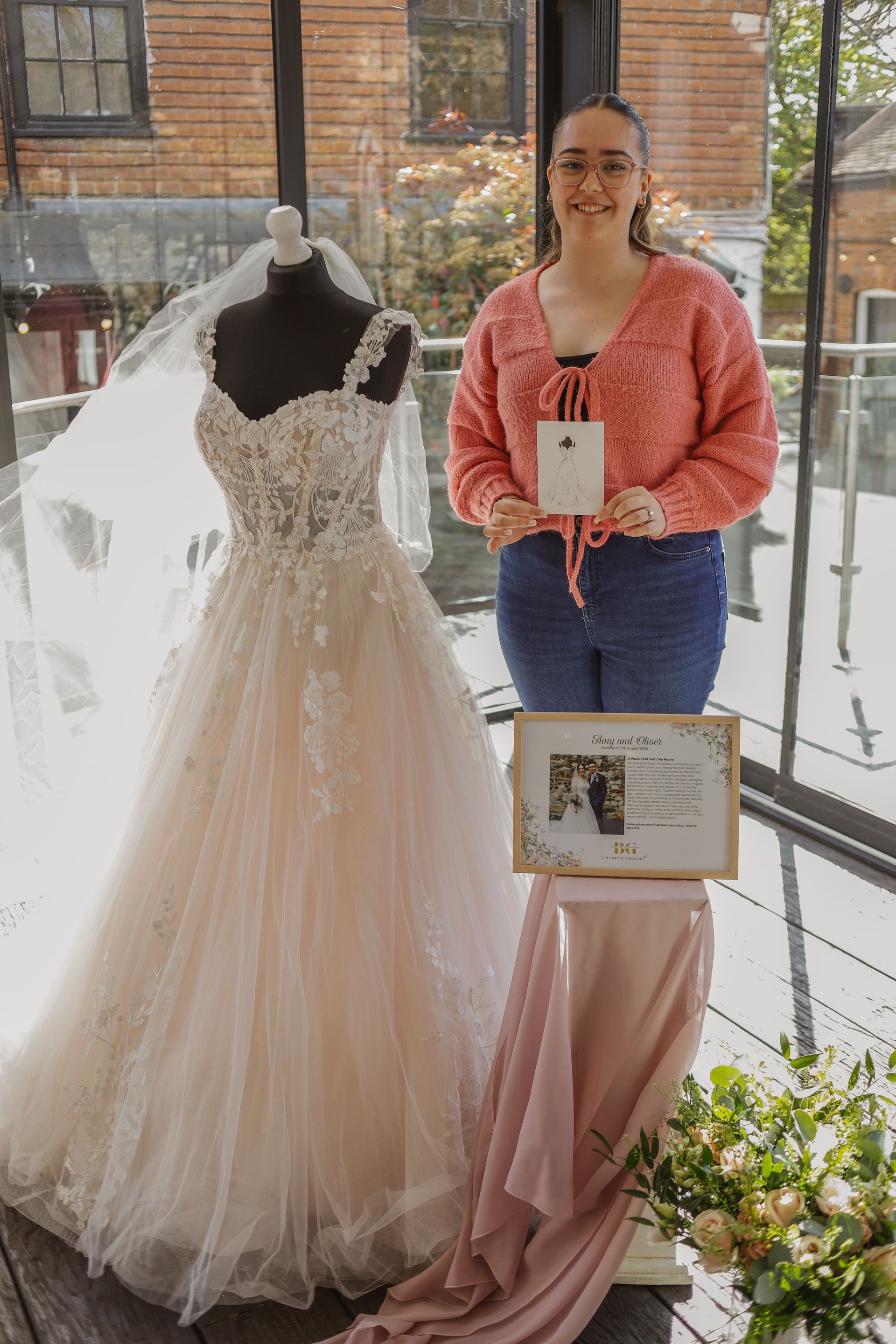 Bridal gown on mannequin beside woman in pink top holding framed sign in a bright showroom