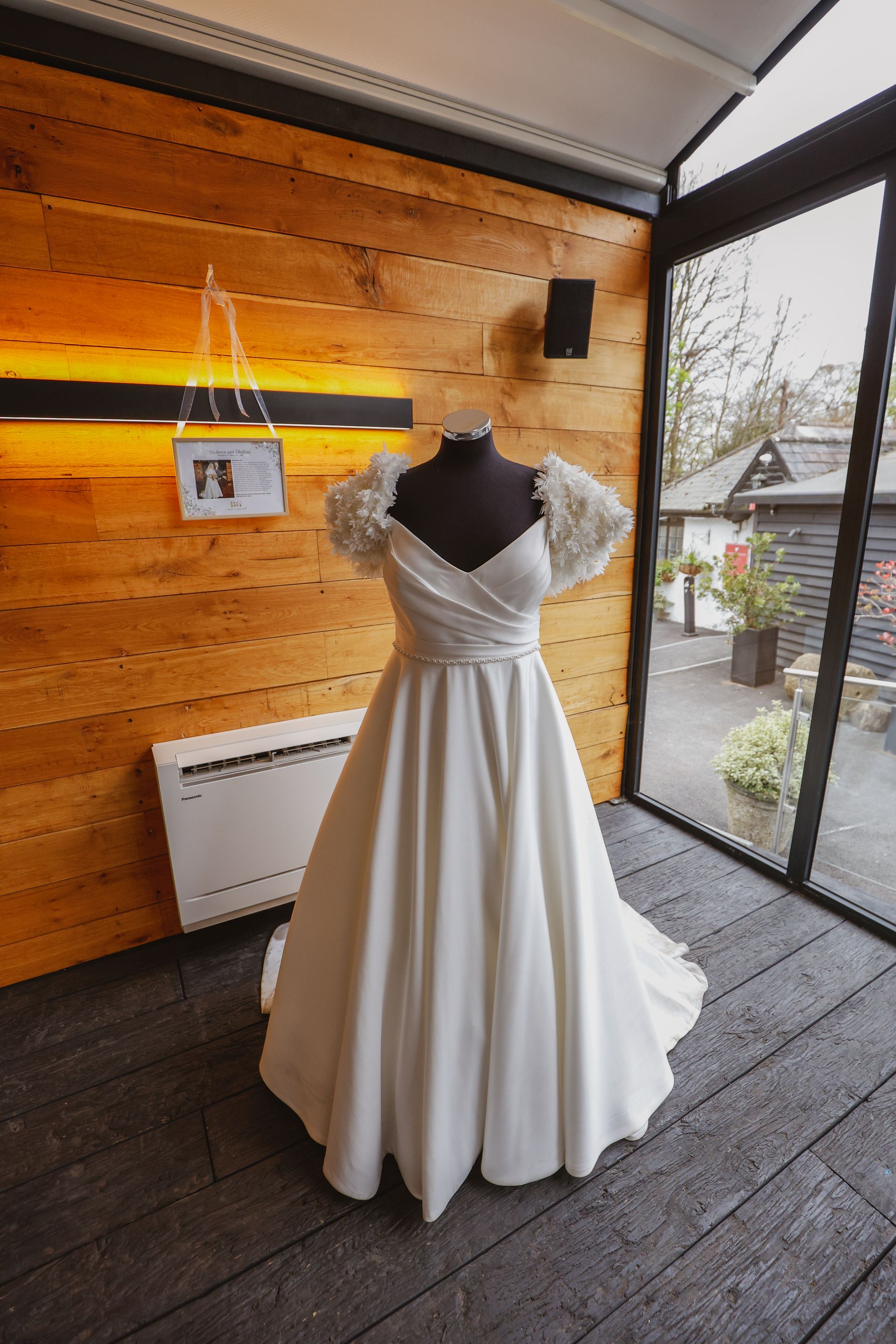 White wedding dress on a black mannequin in a rustic studio with wood walls and large windows.