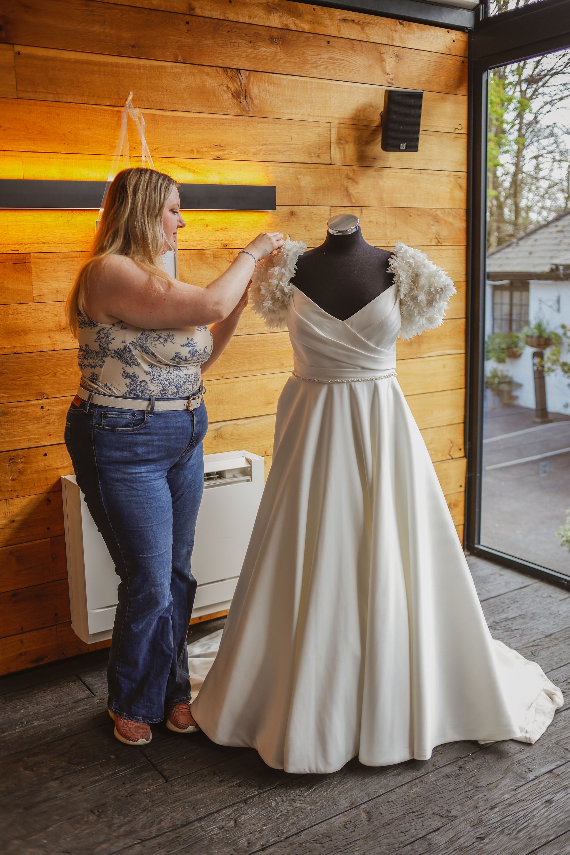 Woman adjusting a white wedding dress on a mannequin in a rustic wood-paneled room