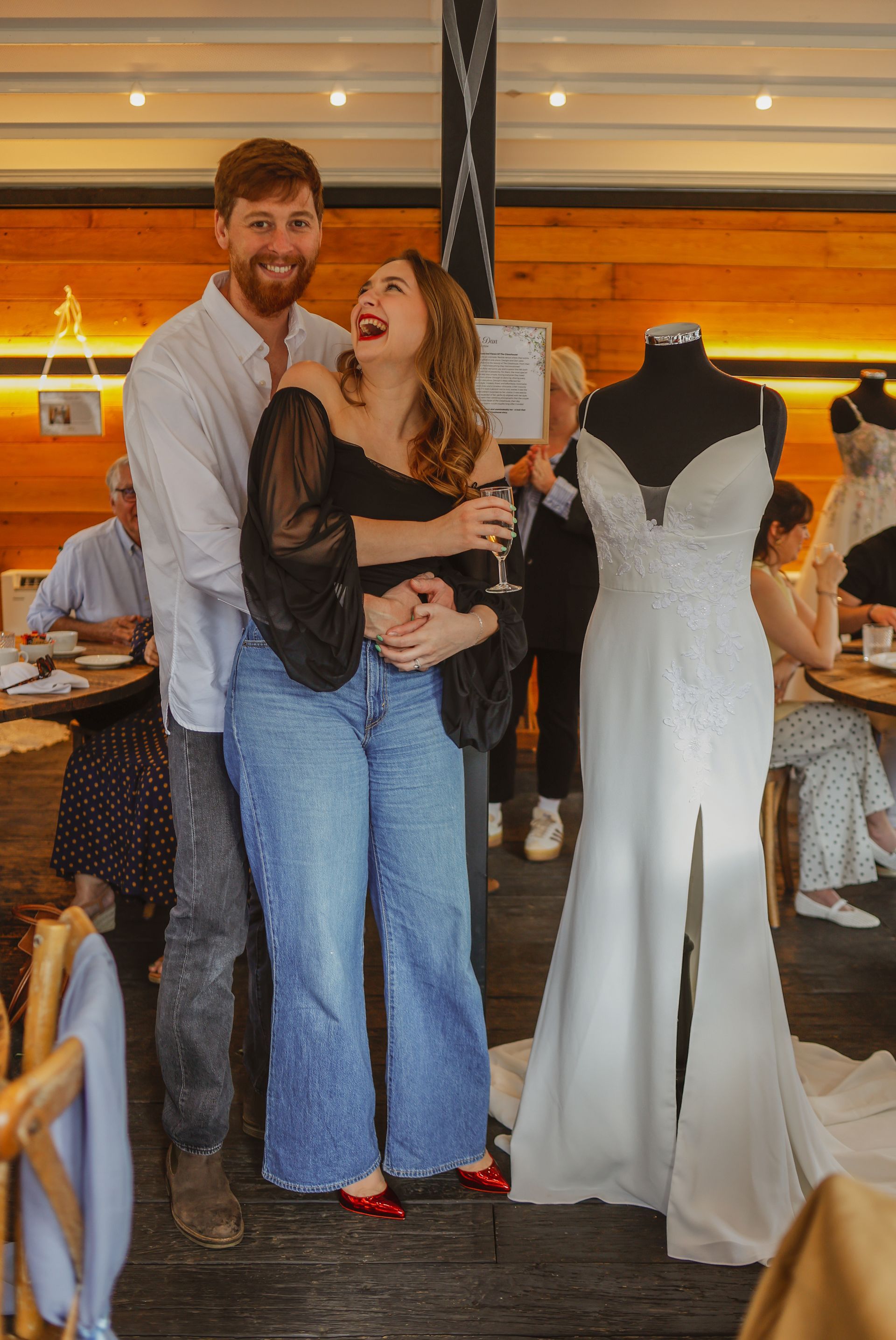 Couple posing indoors beside a white wedding dress on a mannequin, smiling in a warmly lit shop.