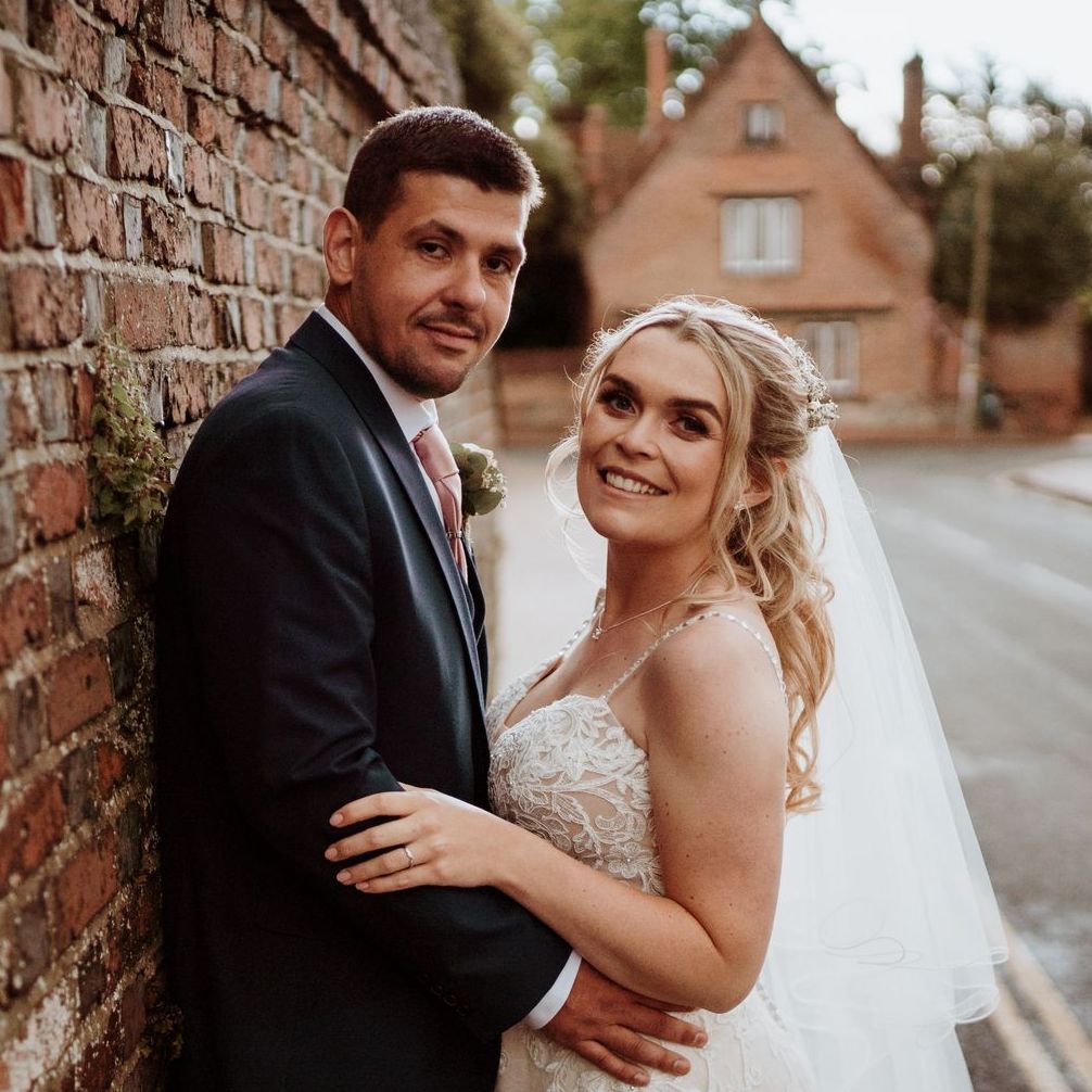 Bride and groom posing by a brick wall on a street, smiling at the camera