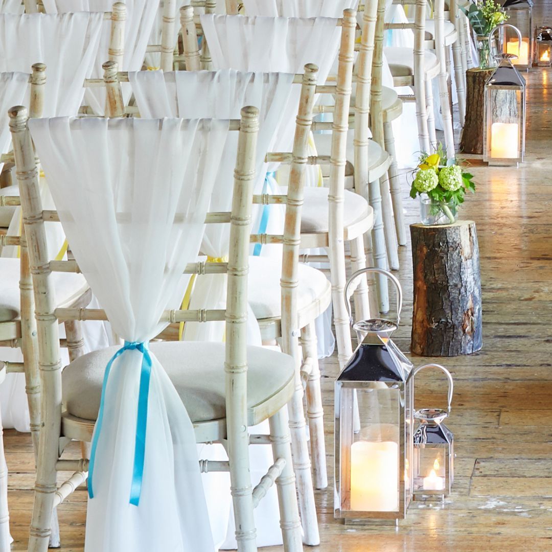 White chiavari chairs with sheer bows and lanterns line a rustic indoor wedding aisle.