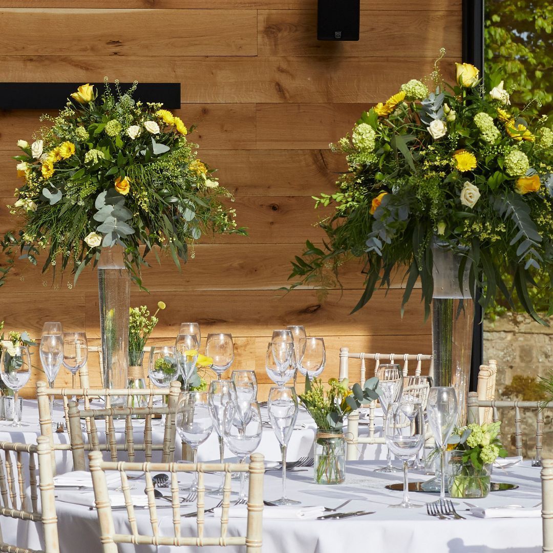 Elegant banquet table with white linens and tall yellow-and-white floral centerpieces in a rustic wooden venue