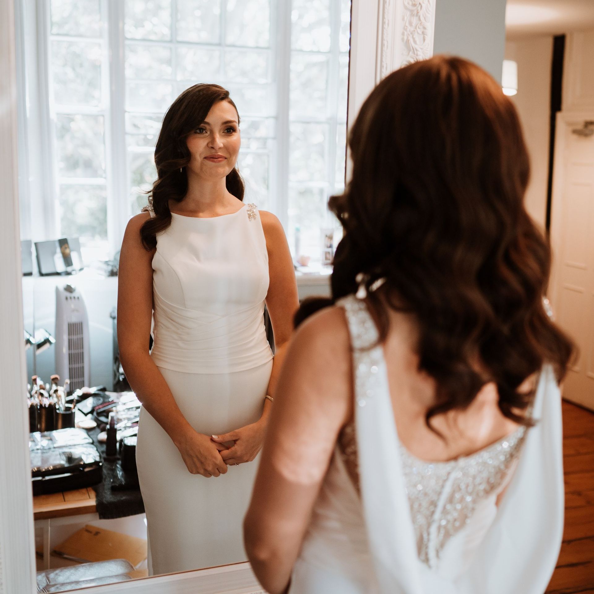 Woman in white dress looking in a mirror, adjusting her wedding gown in a bright room.