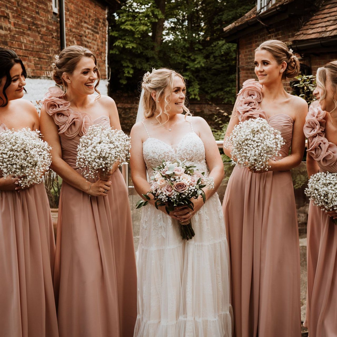 Bridesmaids in blush dresses stand with a bride in a white gown holding bouquets outdoors.