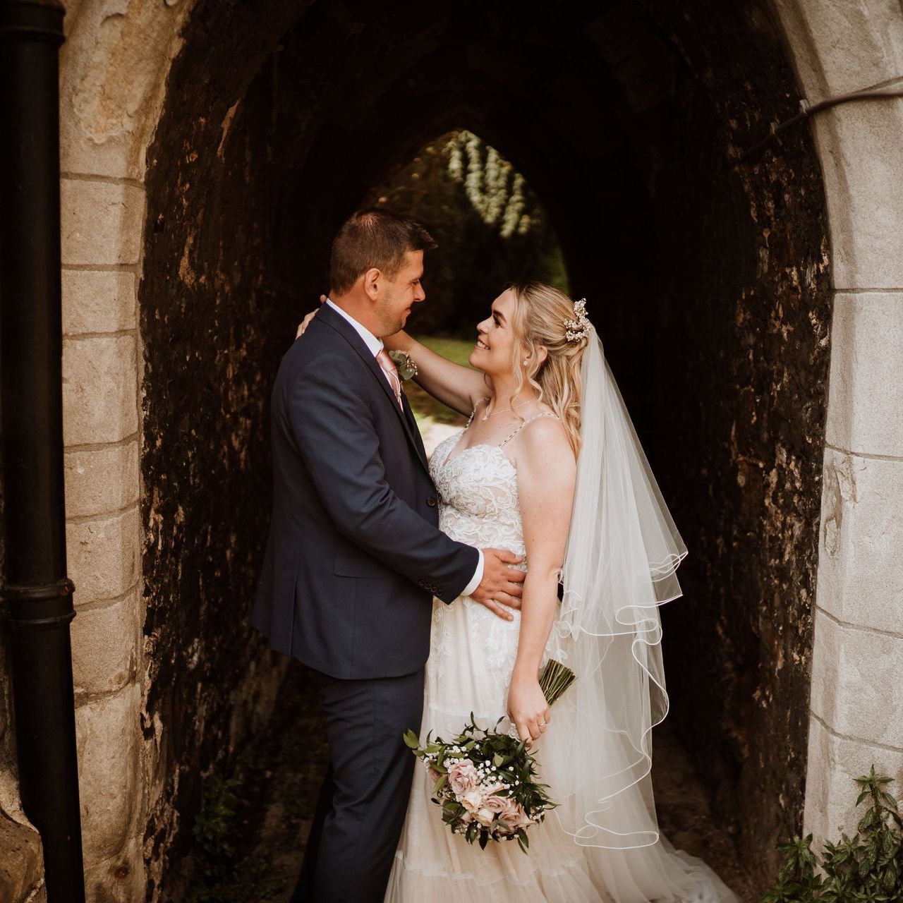 Bride and groom embracing in a stone archway, sharing a tender moment in wedding attire.