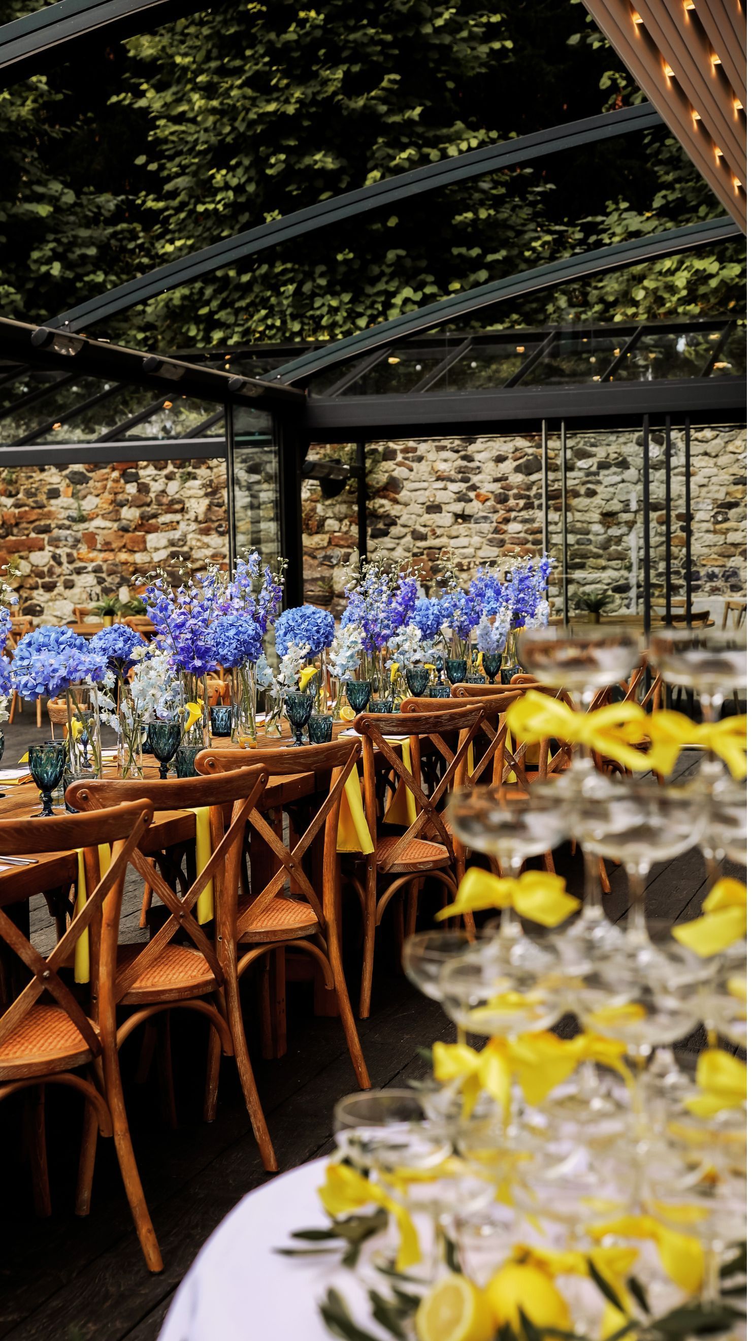 Elegant outdoor dining table with yellow glassware and blue flowers under a pergola