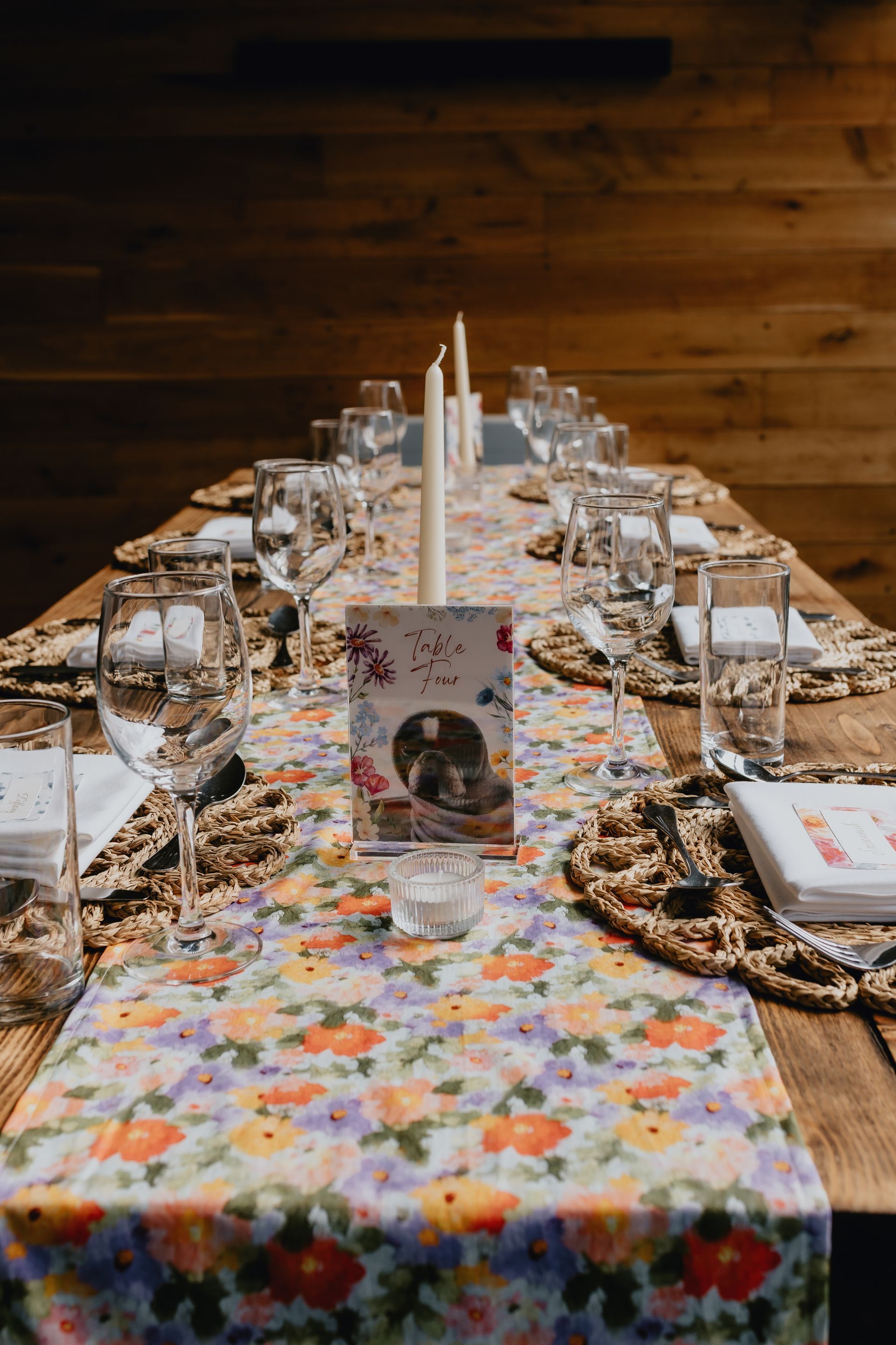 Decorated long dining table with floral runner, glasses, plates, and candles in a rustic room