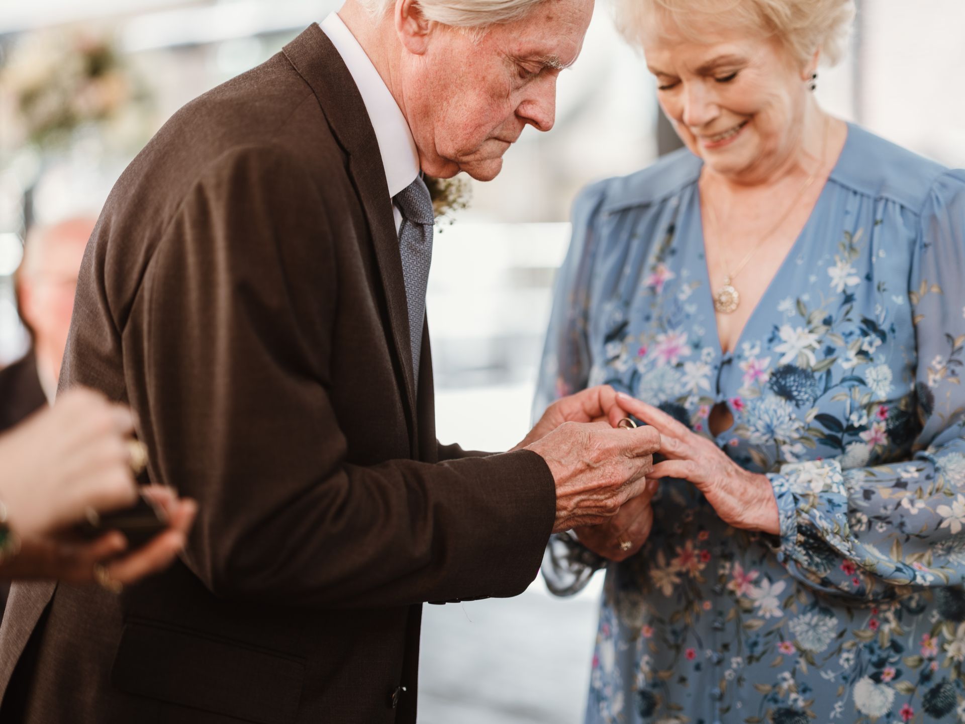 Elderly couple exchanging rings at a formal ceremony, with a blue floral dress and brown suit.