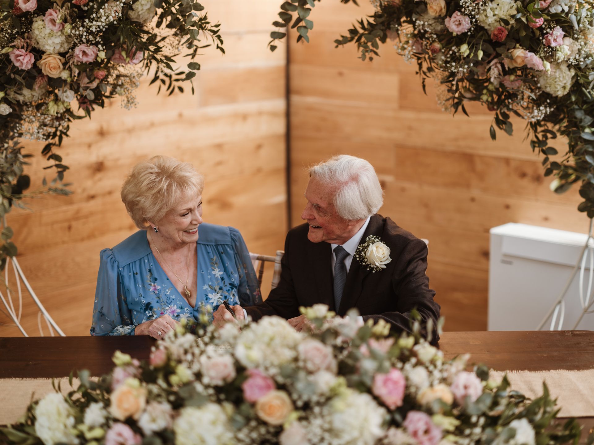 Elderly couple seated at a floral-decorated wedding table, smiling at each other beneath an arch of flowers