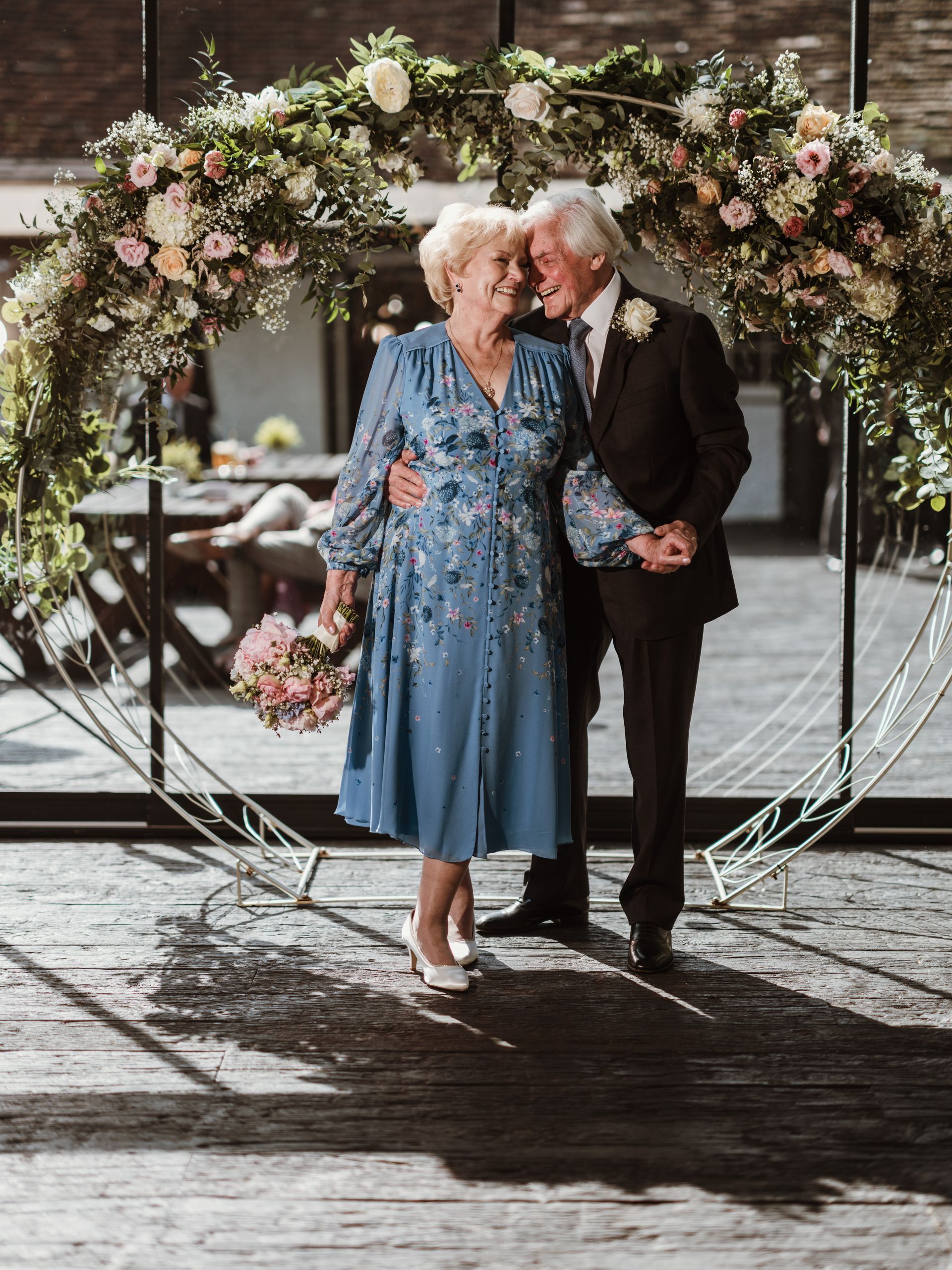 Two people holding hands beneath a flower-covered arch, dressed in blue and black, outdoors on a wet floor