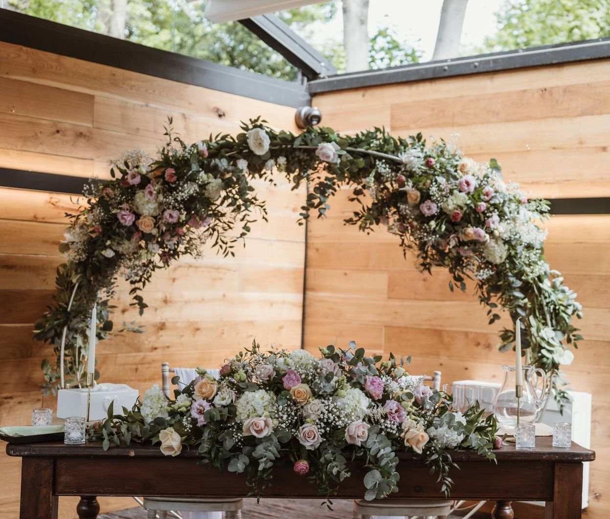 Floral wedding arch with pink and white flowers on a wooden table in a rustic indoor setting