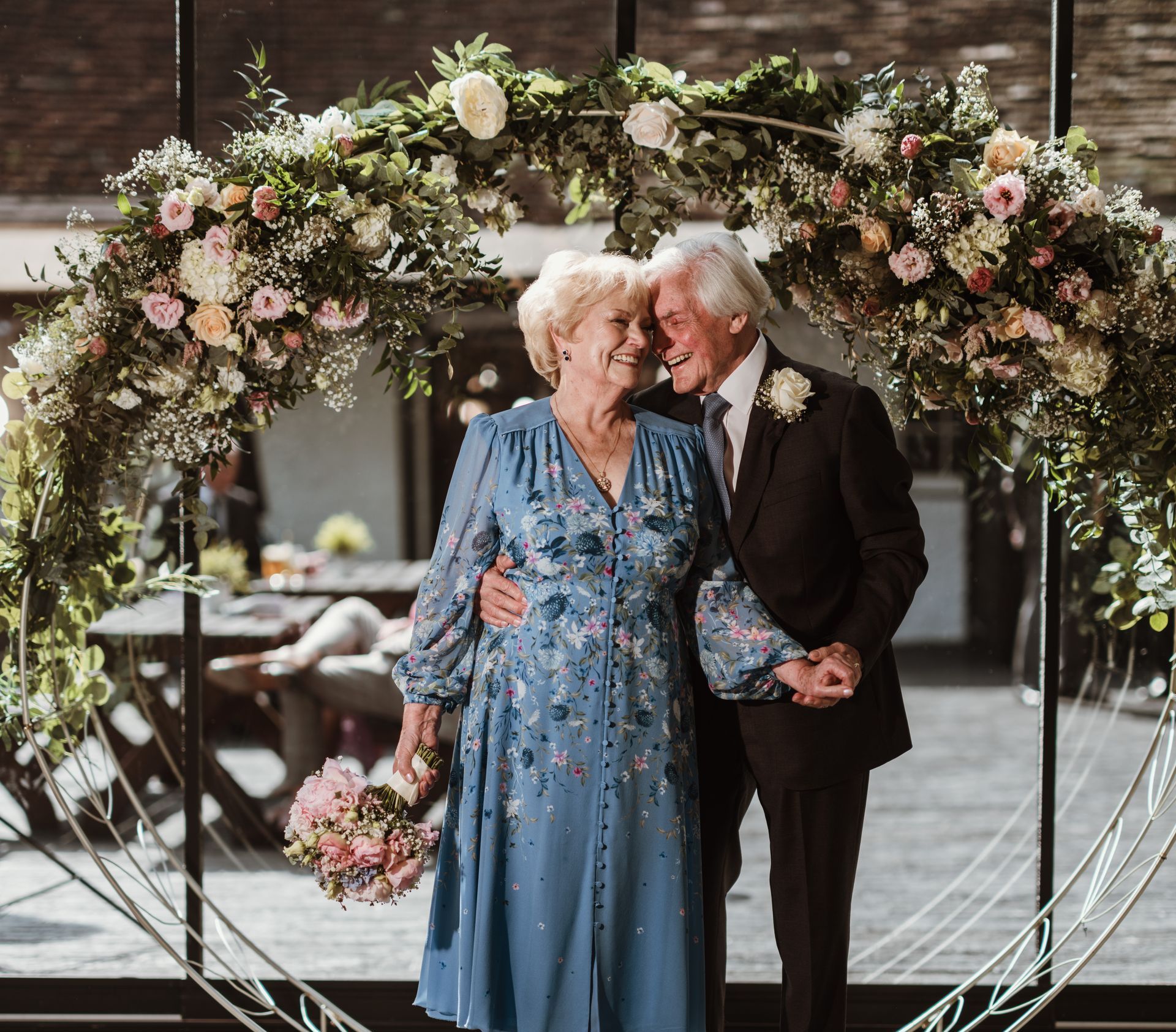 Elderly couple smiling under a floral arch, dressed in blue and black, embracing at an indoor celebration