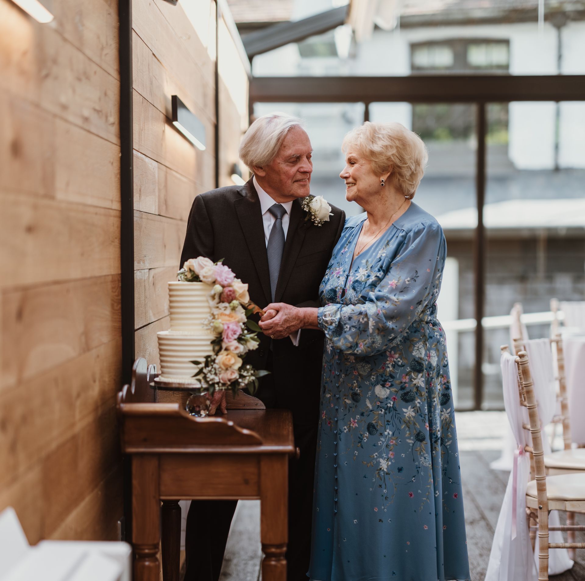 An elderly couple holding hands at an indoor wedding reception, dressed in formal attire beside a cake table
