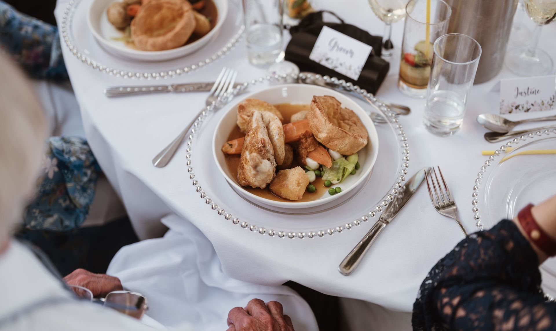 A plated dinner on a white tablecloth with bread, cutlery, wine glasses, and guests’ hands around the table