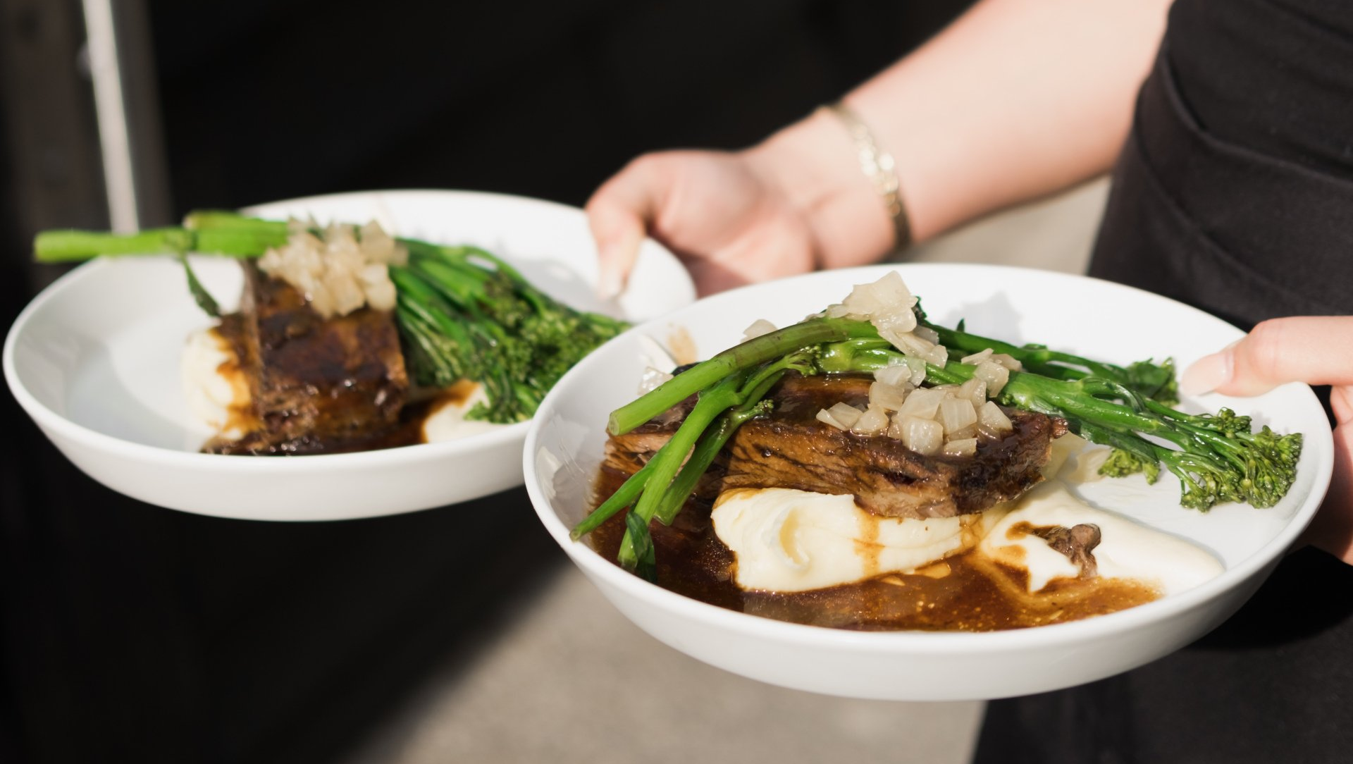 Two bowls of white rice topped with braised meat and greens, held by hands